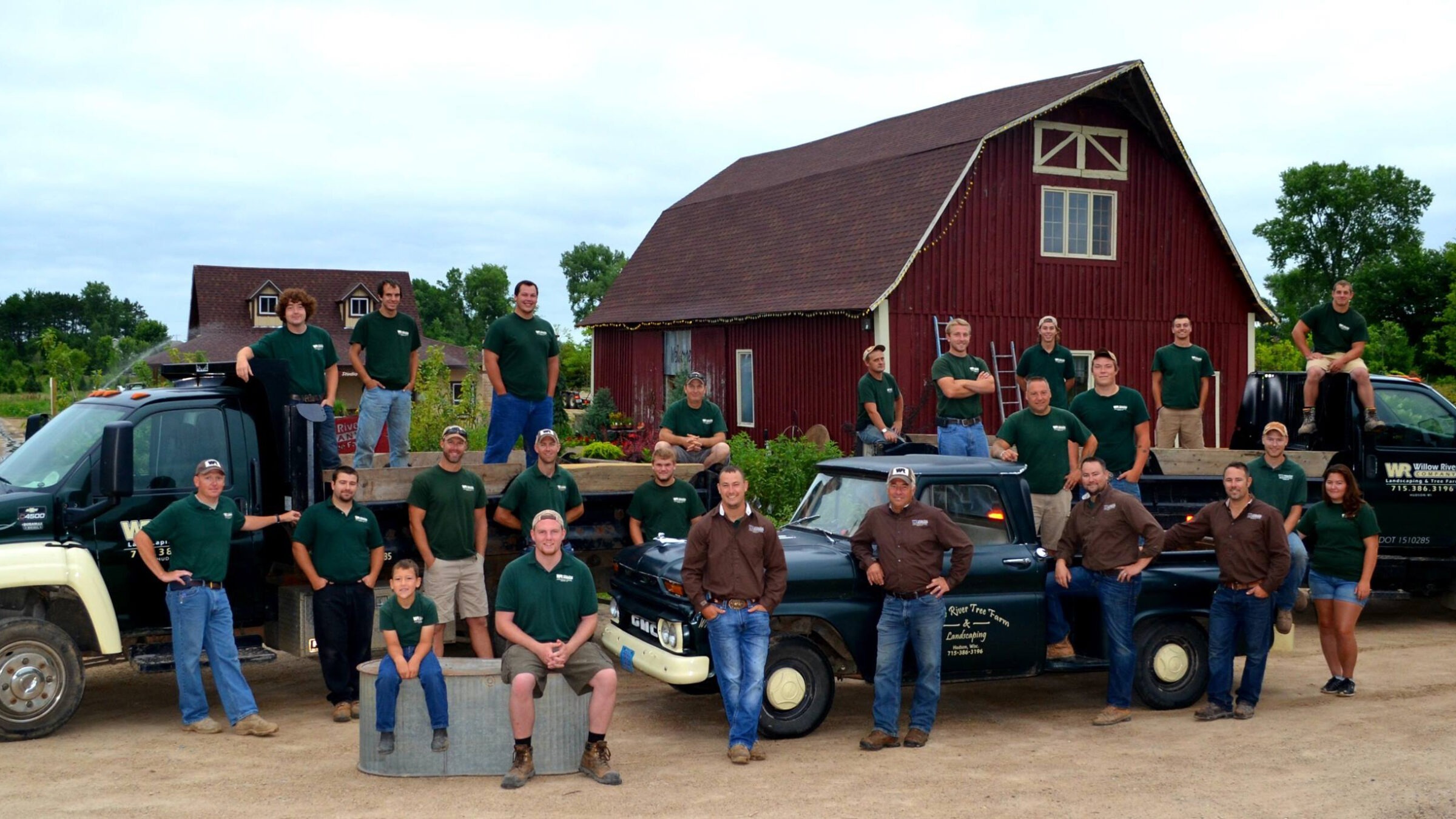 A group of people in matching green shirts pose with trucks in front of a large red barn, surrounded by greenery.