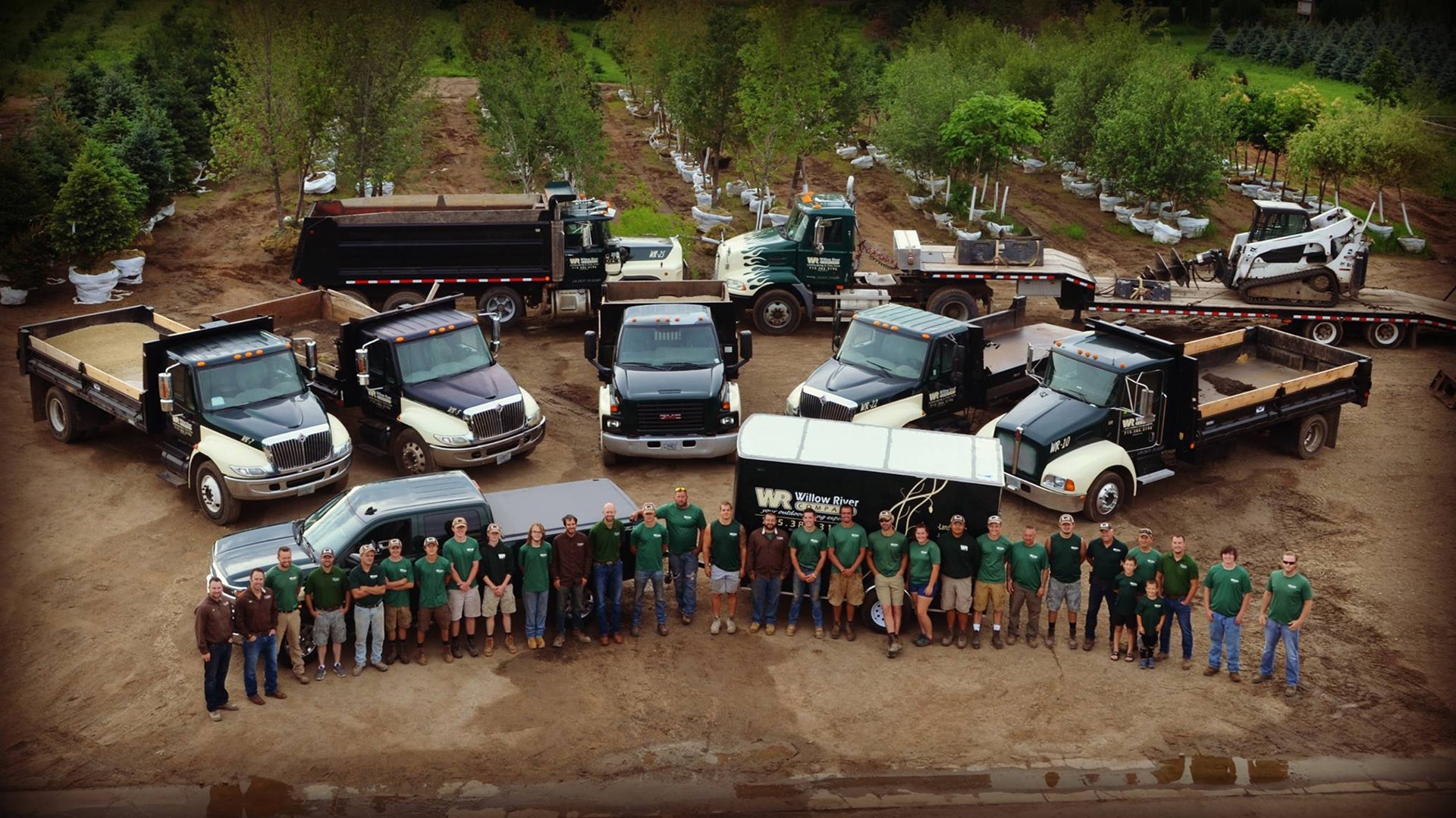 A group of people wearing green shirts stand in front of various work trucks and equipment, surrounded by trees in a nursery setting.