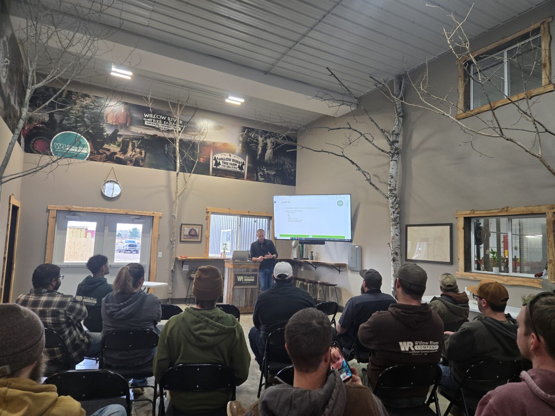 Indoor meeting with seated attendees facing a screen displaying an agenda. A person stands at a podium in a rustic-themed setting.