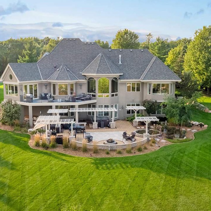 Elegant two-story house with large windows, surrounded by lush green lawn and trees, featuring spacious outdoor patio with pergolas and furniture.