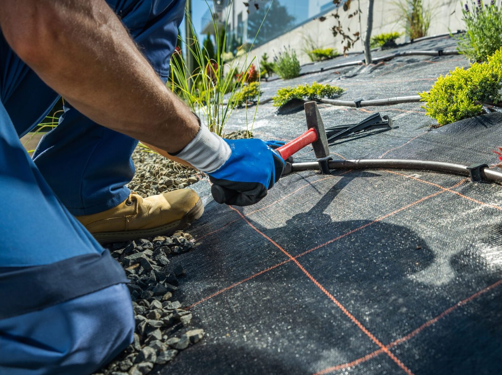 A person installs irrigation on a landscaped slope, using a hammer and gloves, surrounded by plants and gravel, beneath a sunny sky.
