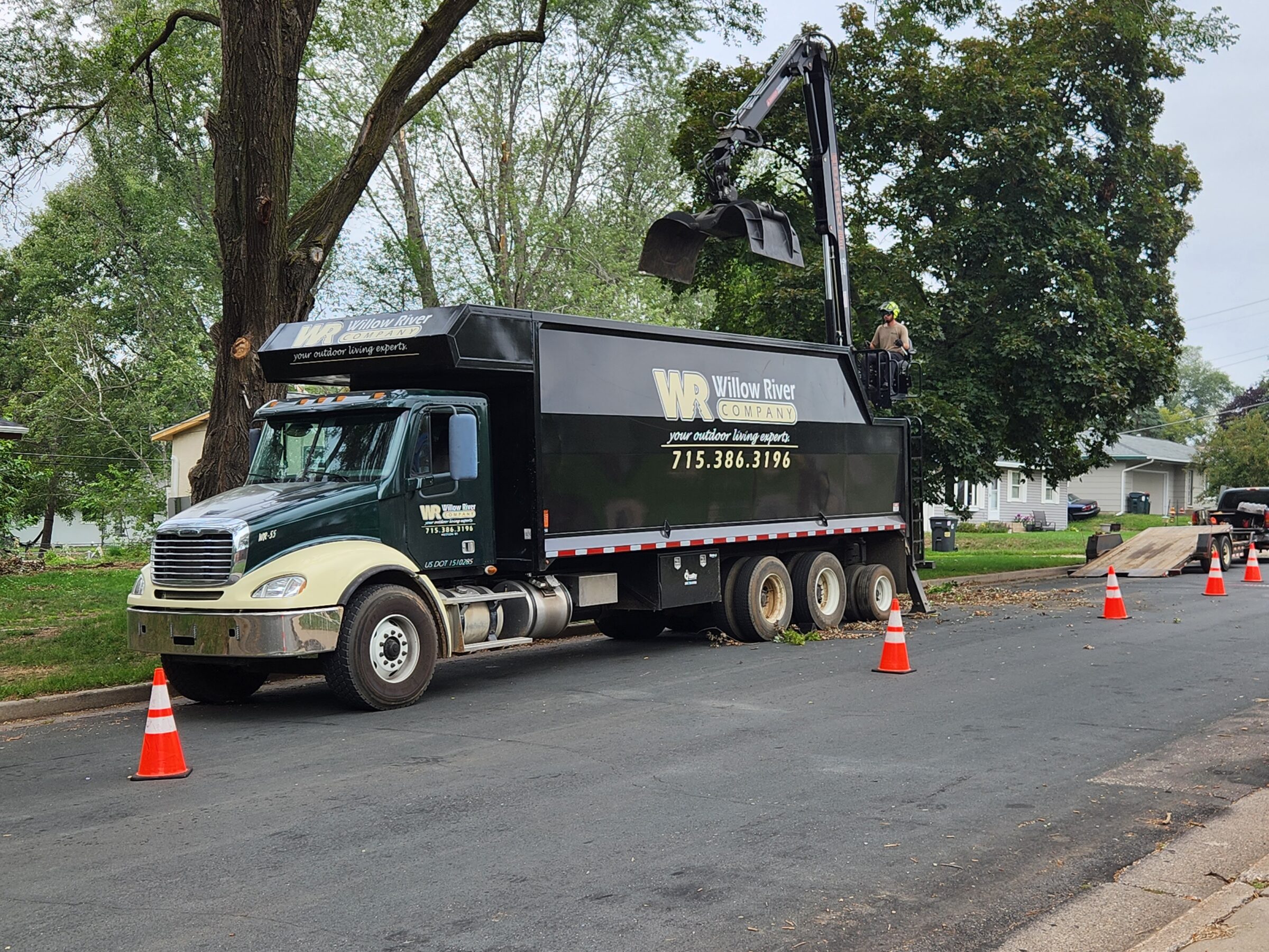 A truck from Willow River Company conducts tree work on a residential street, with cones marking the area and a person managing equipment.