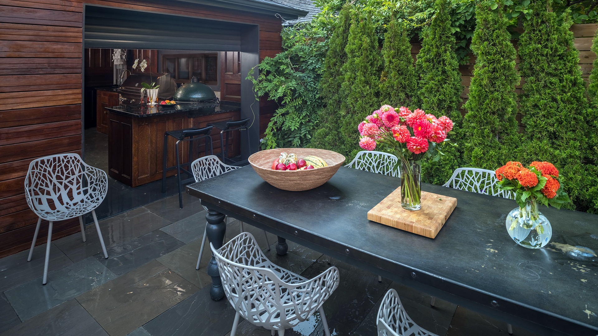 Outdoor patio with modern chairs, a table adorned with vibrant flowers and fruit bowl, adjacent to an open kitchen with a grill.