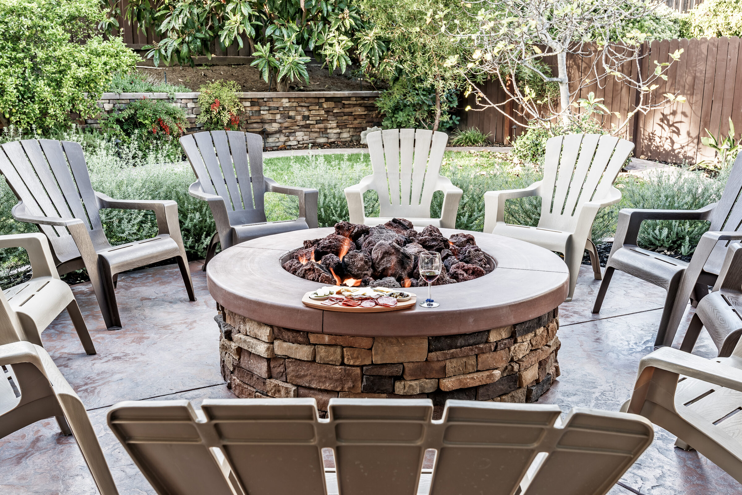 Circular stone fire pit surrounded by chairs in a garden setting; platter with snacks and a glass of wine on the edge.