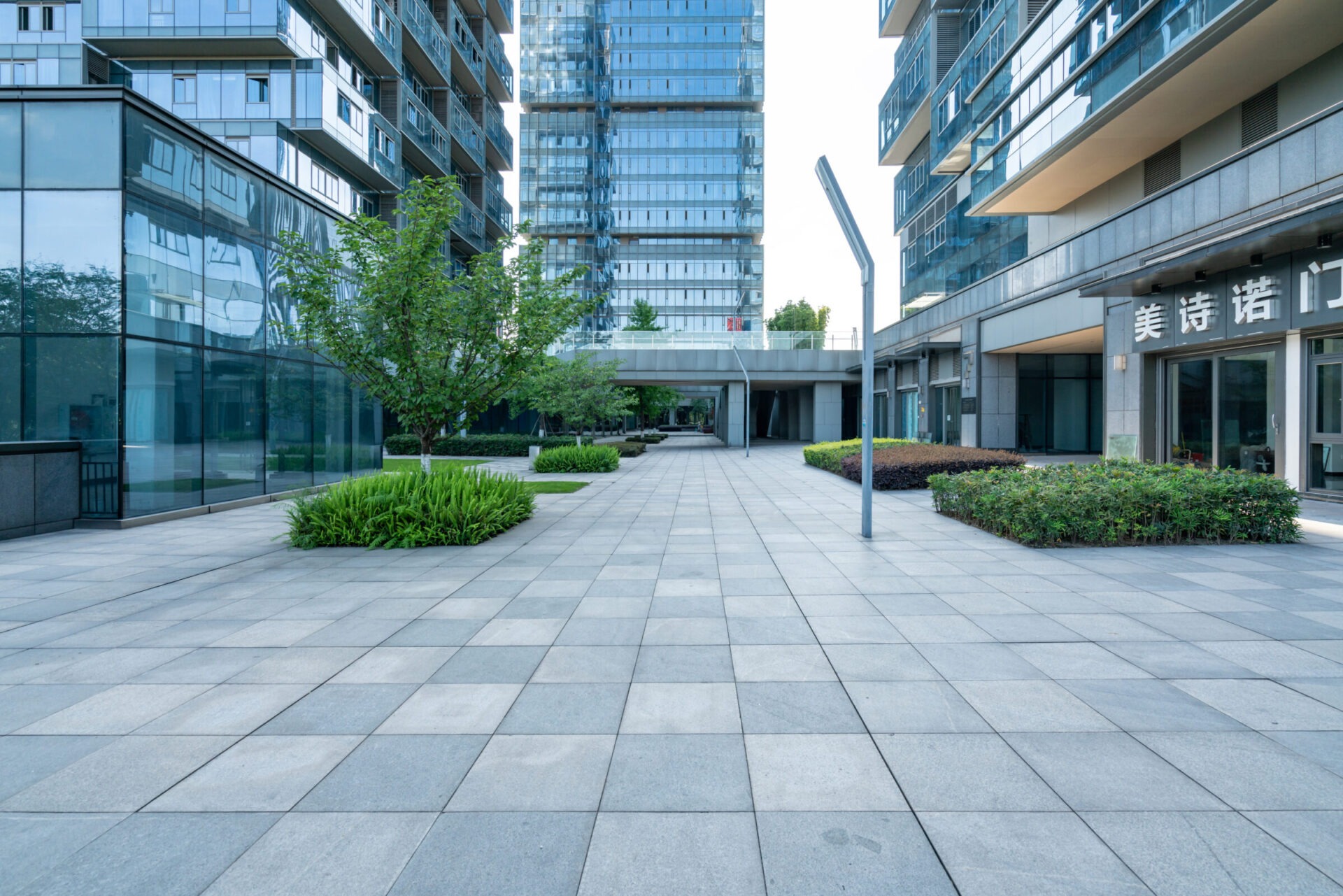 Modern courtyard with glass buildings, trees, and paved walkway. Tall structures surround the area, creating a calm and orderly urban landscape.