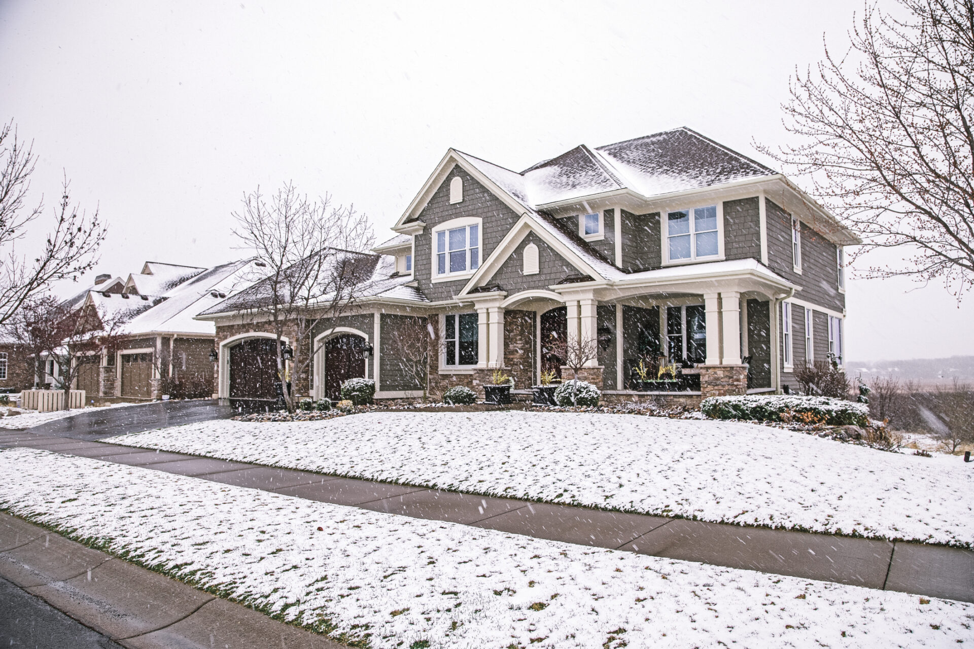 Snow-covered suburban house and driveway, surrounded by bare trees and bushes, under a gray sky, indicating a winter setting.