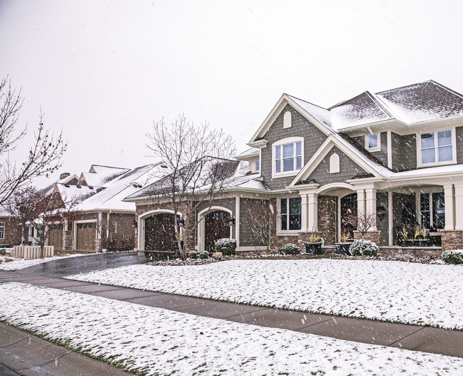 Snow-covered suburban house and driveway, surrounded by bare trees and bushes, under a gray sky, indicating a winter setting.