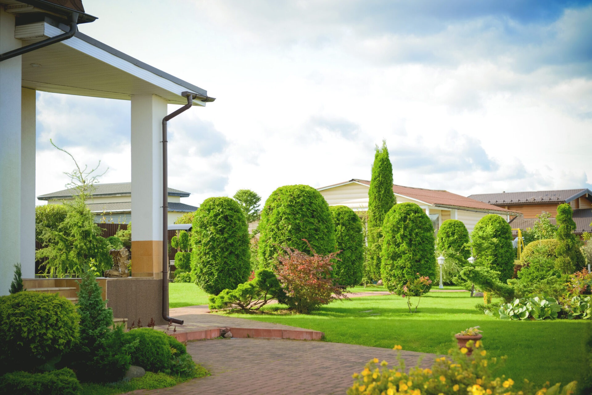 A well-manicured garden with neatly trimmed shrubs, surrounded by residential homes under a partly cloudy sky. No landmarks or people are visible.
