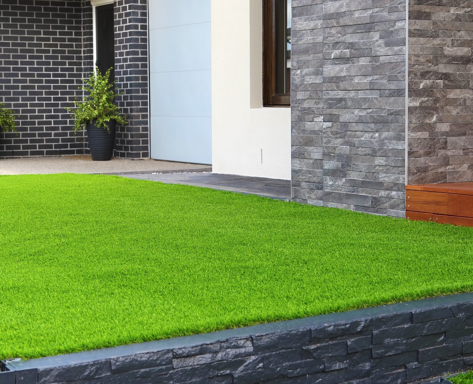 Modern house exterior with gray brick walls, artificial grass lawn, and two planters. The design features sleek lines and contrasting textures.