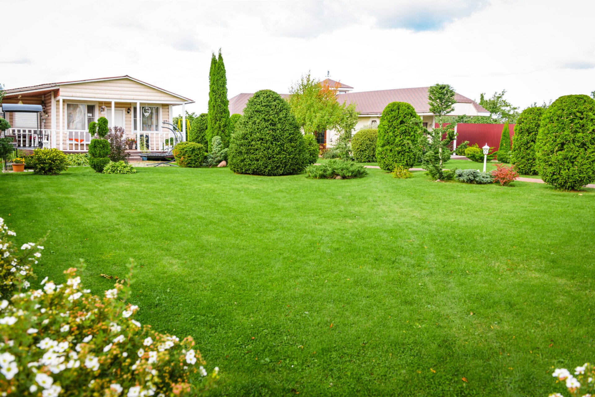 A landscaped garden with neatly trimmed bushes and lush green lawn surrounds a cozy house with a porch and large windows.