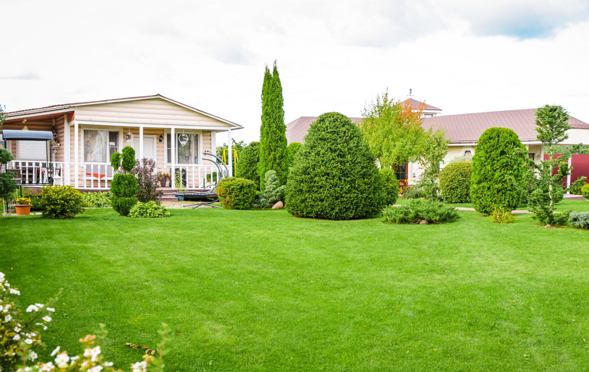 A landscaped garden with neatly trimmed bushes and lush green lawn surrounds a cozy house with a porch and large windows.