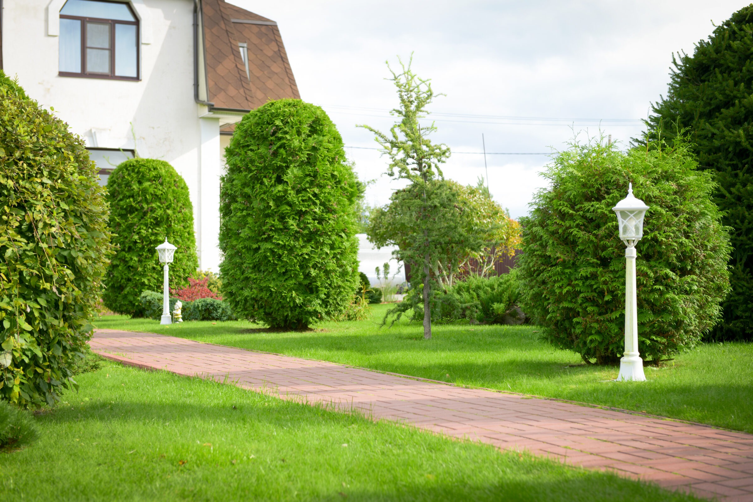 A brick path through a lush garden with trimmed hedges and white lampposts leads to a building with a brown roof.