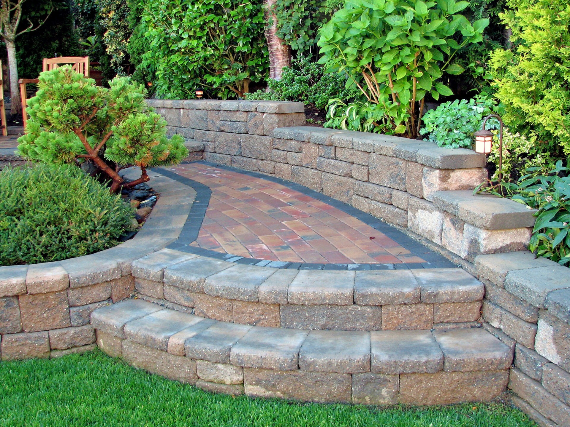A landscaped garden with curved brick pathway, low stone walls, lush greenery, and small trees, creating a serene, inviting outdoor space.