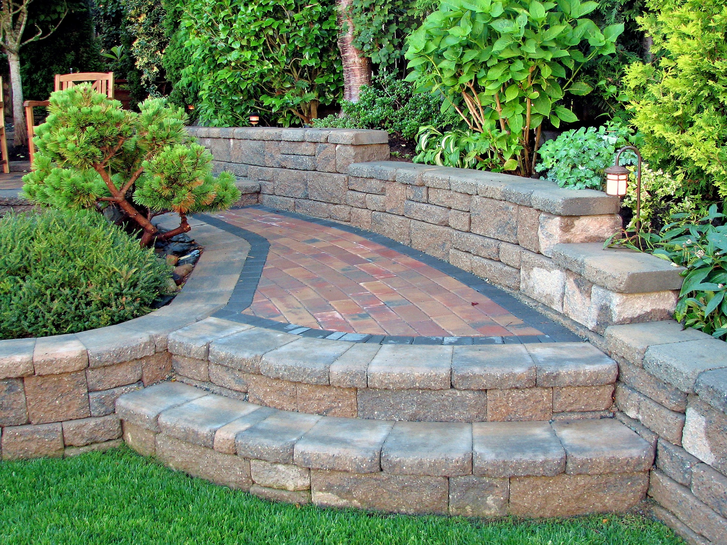 A landscaped garden with curved brick pathway, low stone walls, lush greenery, and small trees, creating a serene, inviting outdoor space.