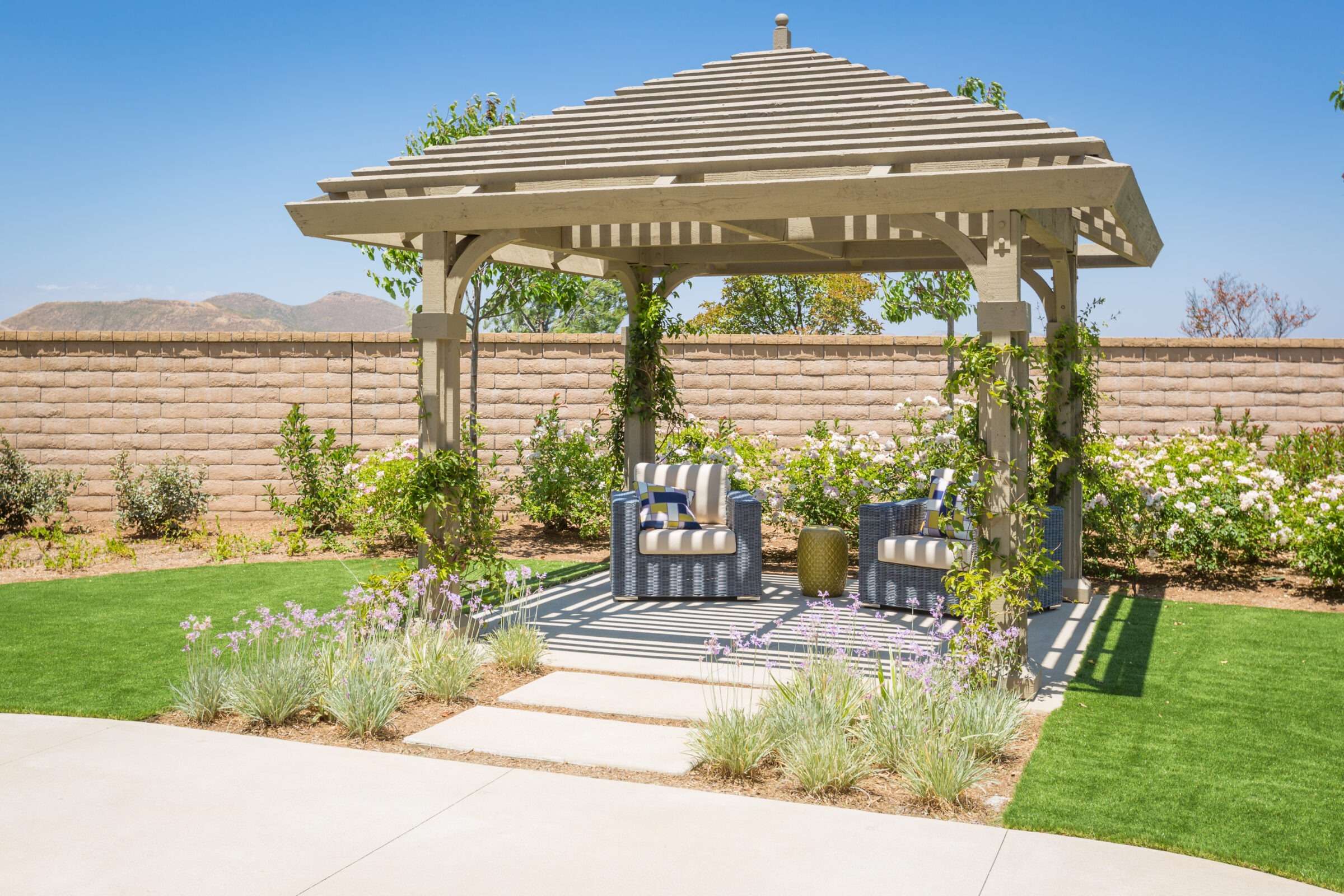 A cozy gazebo with striped chairs and plants in a backyard surrounded by a stone wall, under a clear blue sky.
