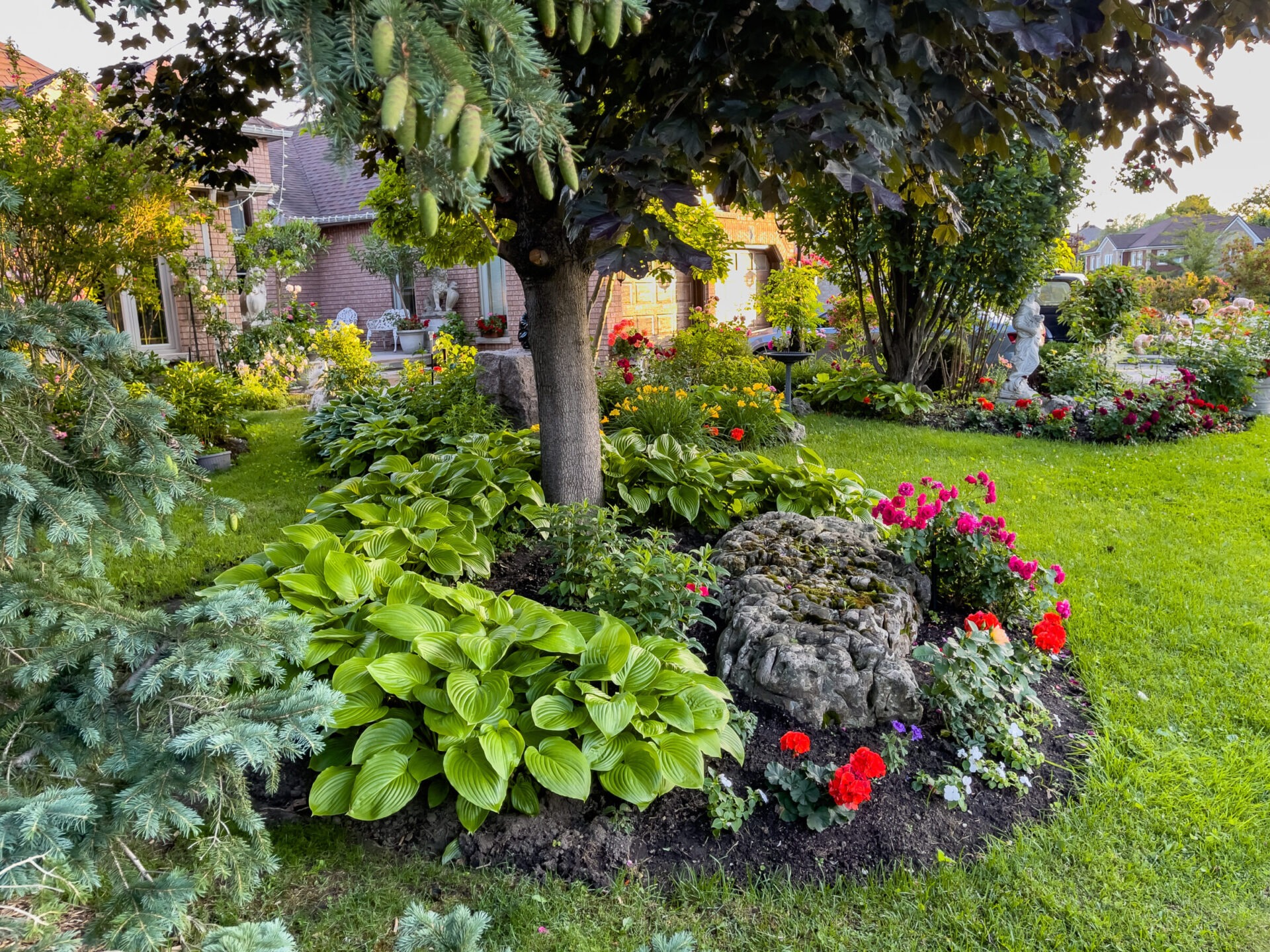 A lush garden with vibrant flowers and various plants surrounds a tree. A person is gardening near a brick house in the background.