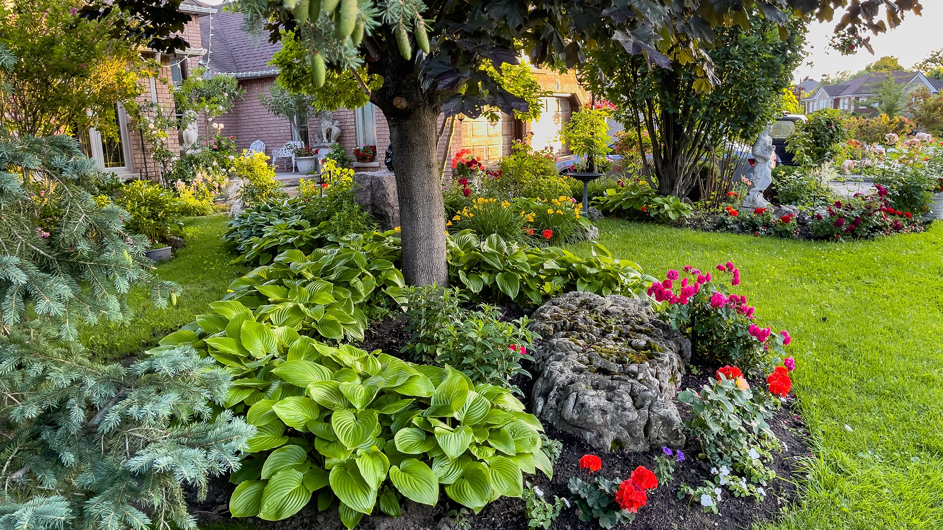 A lush garden with vibrant flowers, a tree, and a statue surrounds a brick house, creating a serene and picturesque setting.