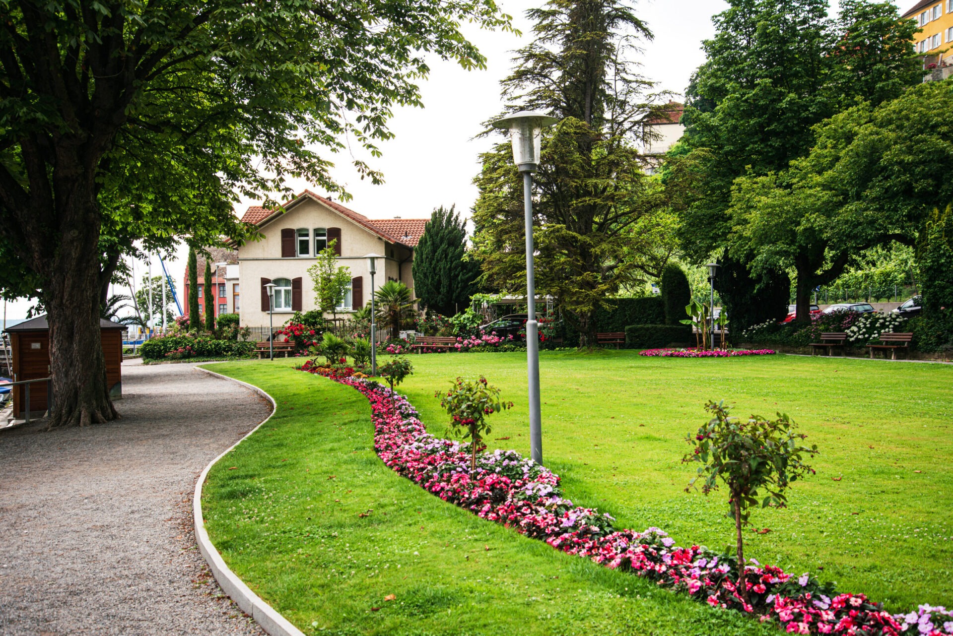 A charming garden with pink flowers leads to a house surrounded by lush trees and greenery, featuring a peaceful, well-maintained pathway.