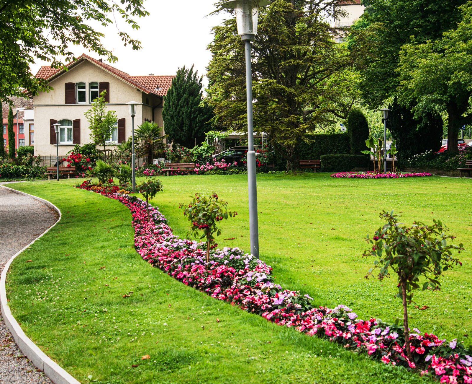 A charming garden with pink flowers leads to a house surrounded by lush trees and greenery, featuring a peaceful, well-maintained pathway.