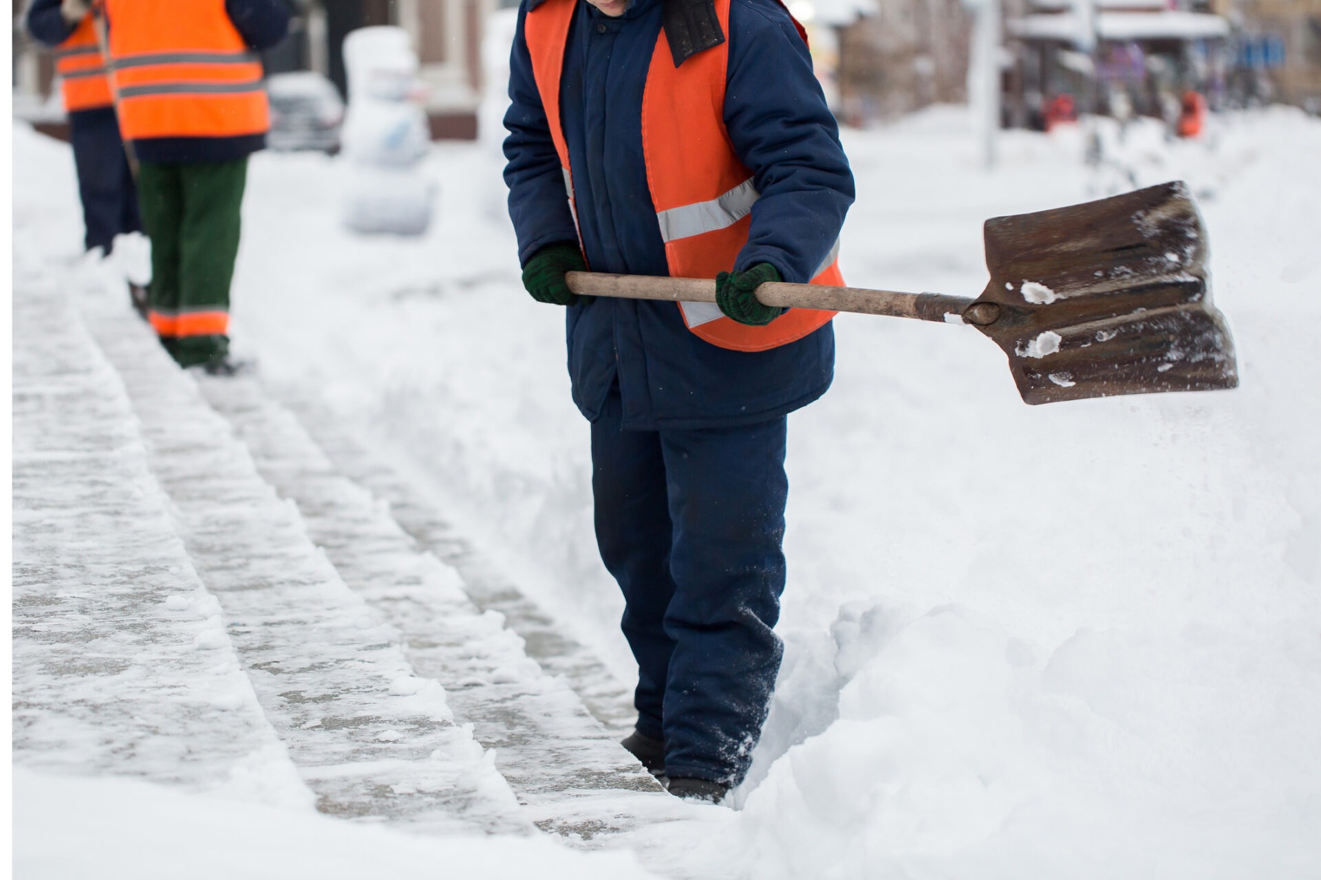 Two persons in orange vests are shoveling snow on a sidewalk. The scene captures winter maintenance work on a snowy day.