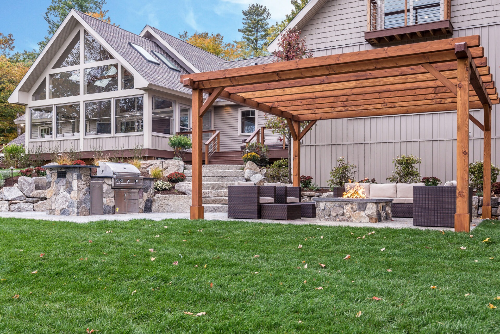 A modern house with large windows and a wooden pergola. Patio has a fire pit, seating, and stone barbecue grill.