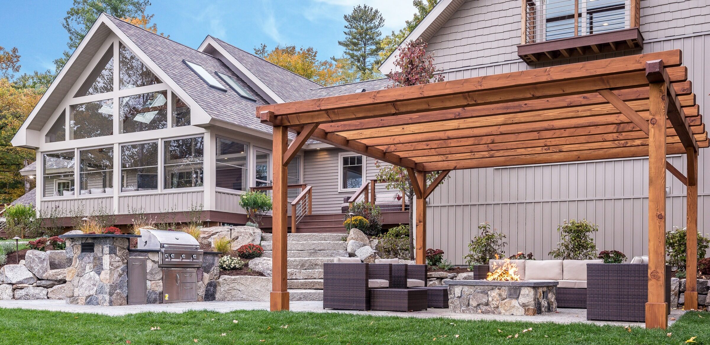Modern house with a pergola, outdoor seating, fire pit, and built-in grill. Surrounded by landscaped garden and trees, under a clear sky.