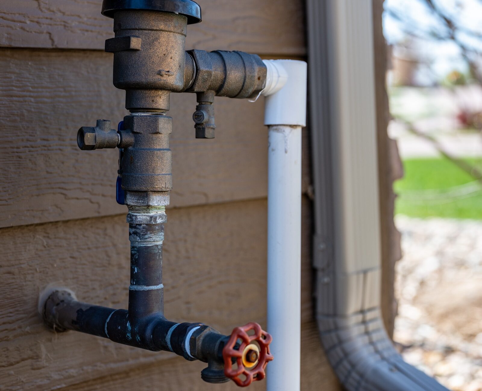 Close-up of a rusty exterior water pipe with valve against a wooden wall. Background shows blurred garden scene with rocks and greenery.