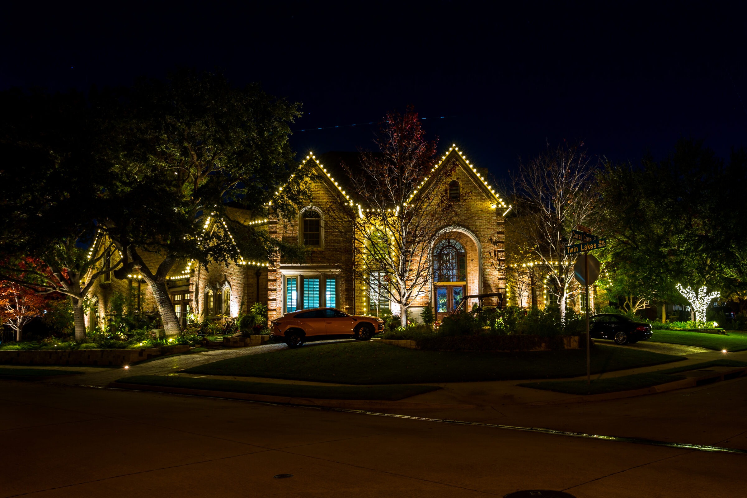 A brick house, illuminated with festive lights at night, is surrounded by trees and parked cars on a peaceful suburban street.