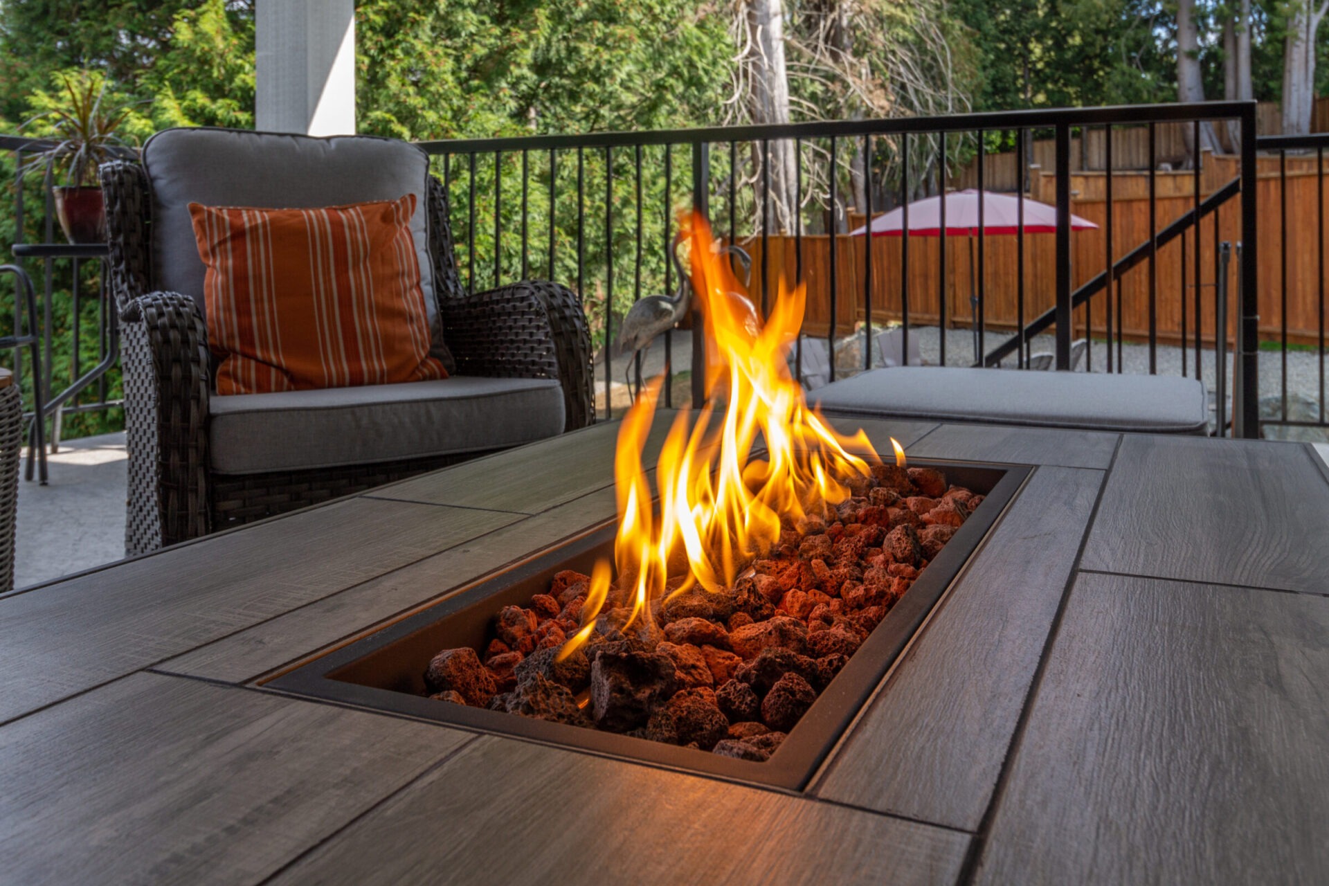 A cozy patio scene with wicker armchairs, a fire pit table, and a red umbrella in a fenced backyard surrounded by trees.