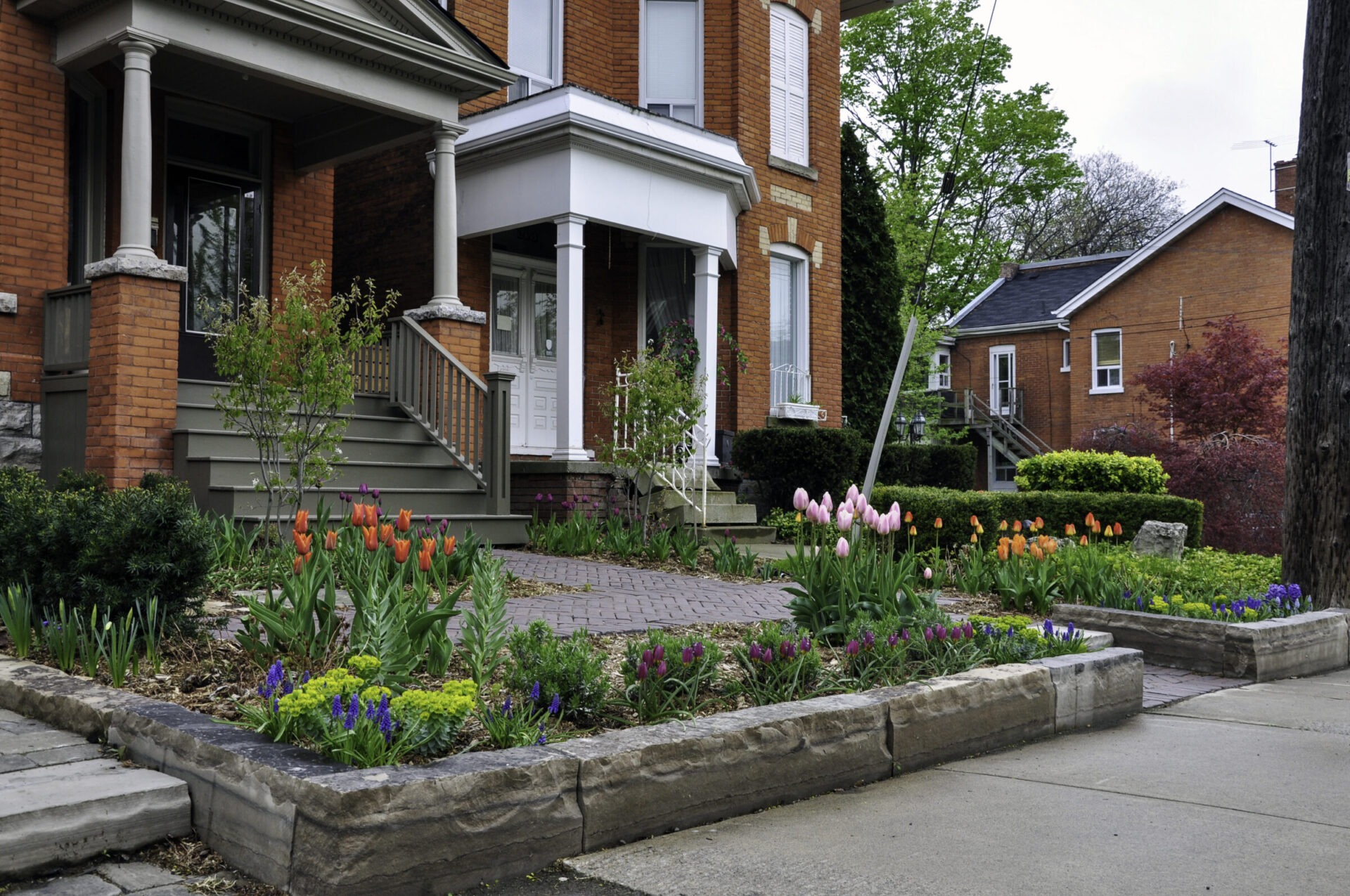 A charming brick house with a well-maintained garden showcasing colorful tulips and vibrant greenery. No recognizable landmarks or historical buildings visible.