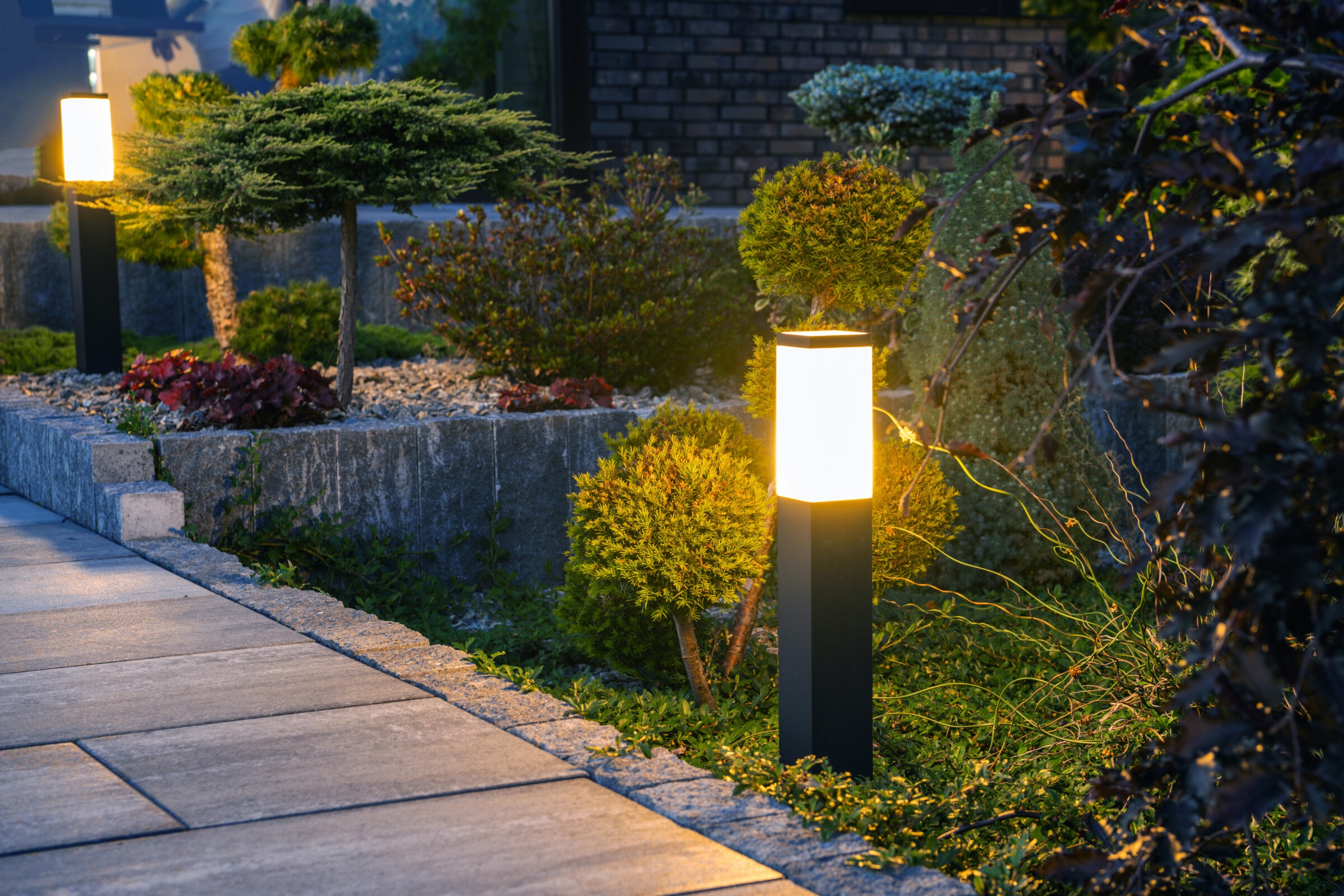 Warmly lit garden pathway with modern stone paving, surrounded by manicured shrubs and soft landscape lighting at dusk.