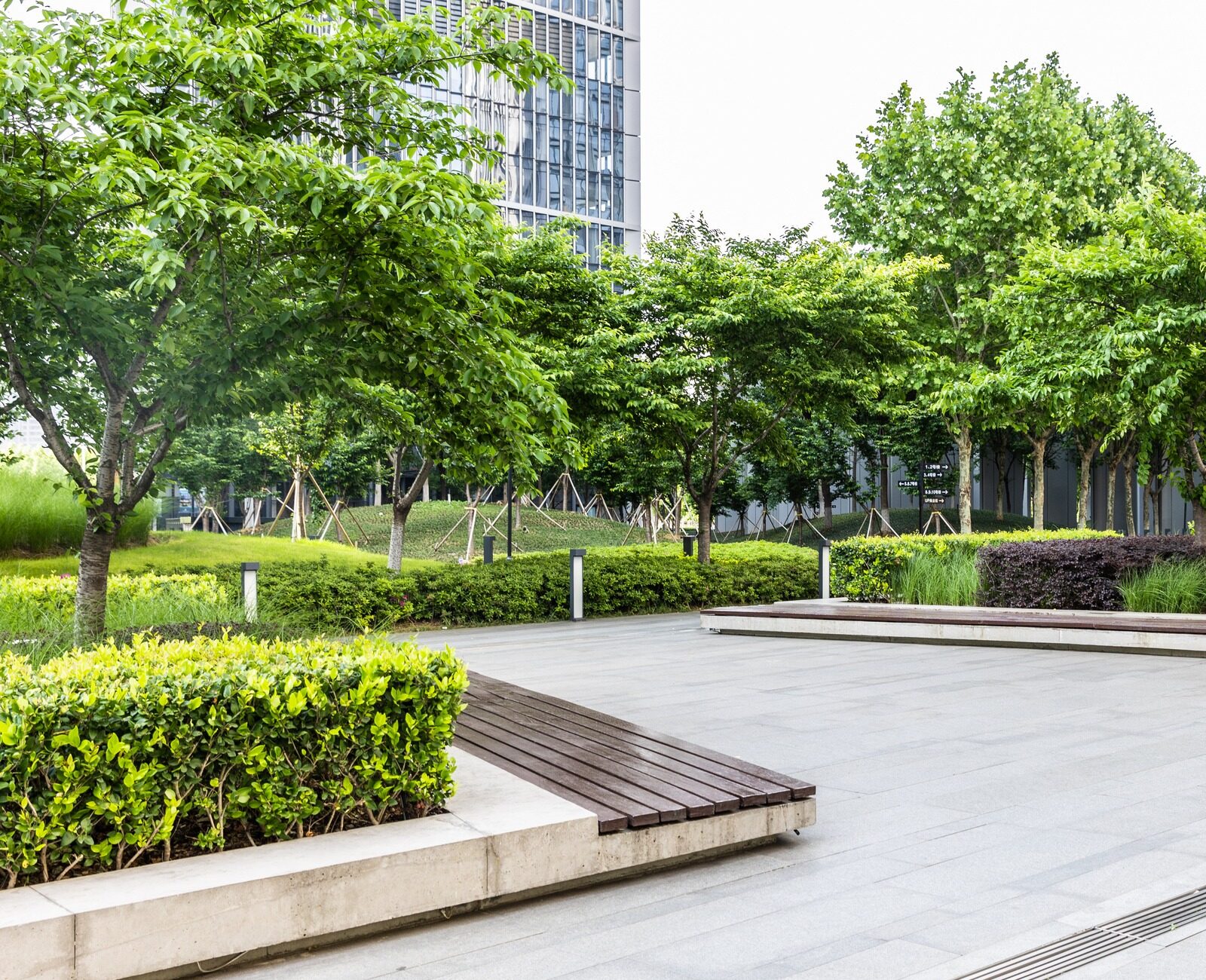 Modern plaza with neatly landscaped greenery and benches. Tall glass buildings surround the area, offering a serene urban oasis amidst the city hustle.