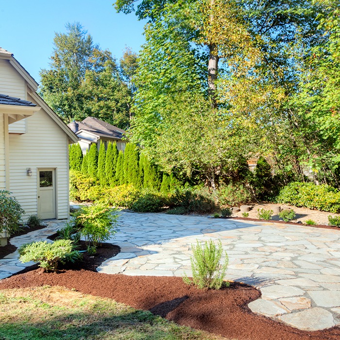 A stone patio beside a house, surrounded by lush green trees and plants, under a clear blue sky in a serene garden setting.