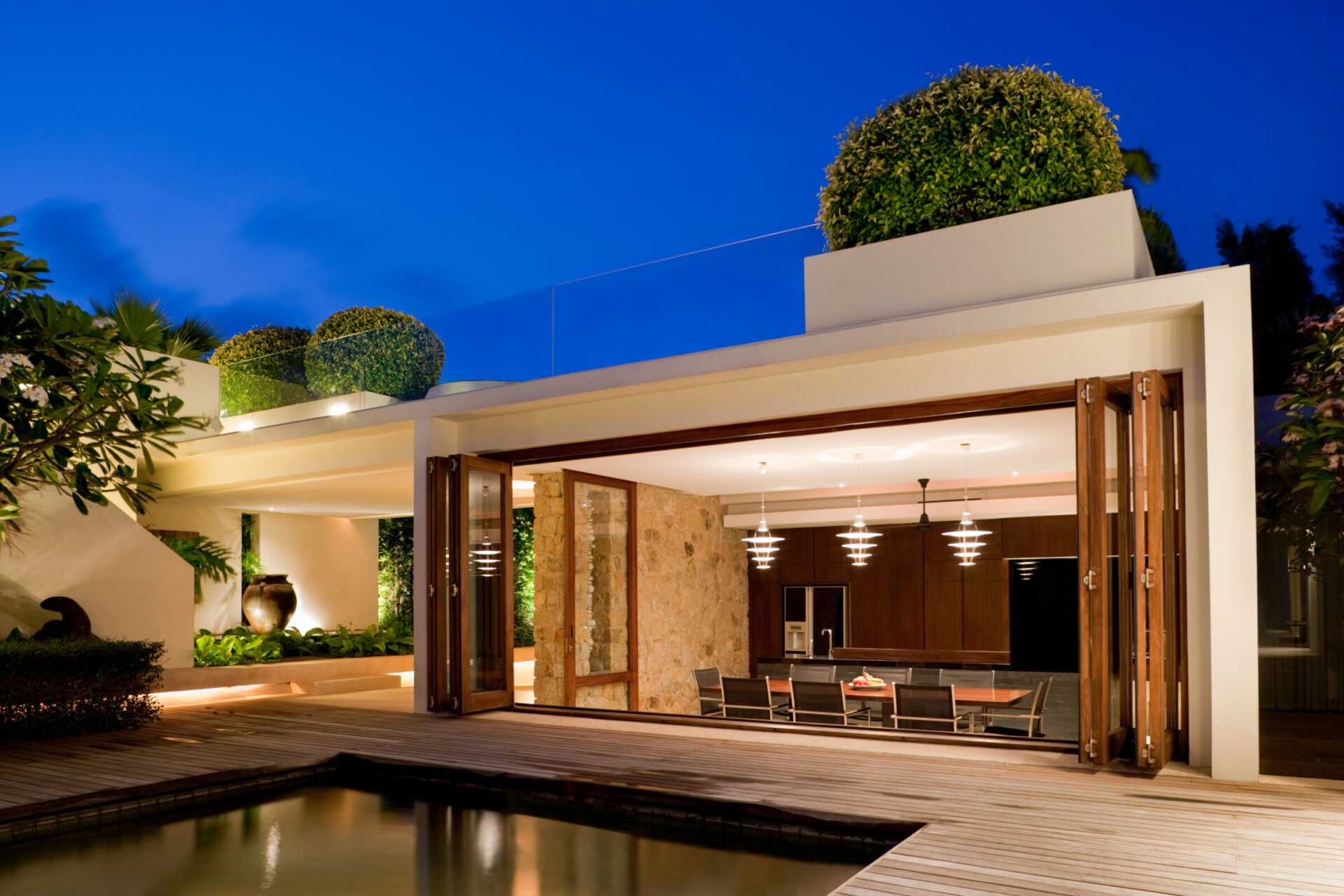 Modern house with open wooden doors leading to a stylish indoor kitchen. Outdoor area features plants and a wooden deck by a pool.