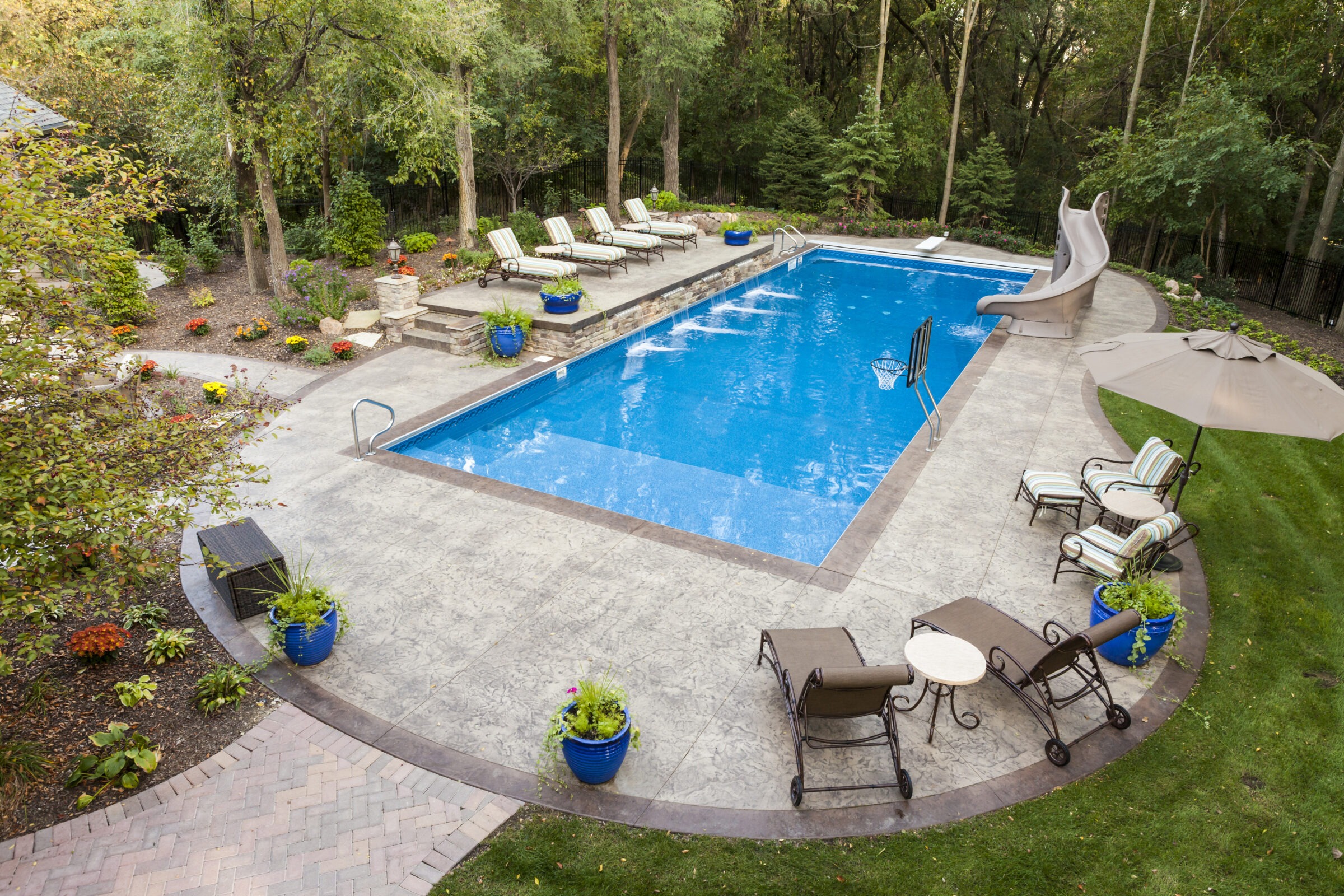 A serene backyard features a large pool with a water slide, surrounded by lounge chairs, potted plants, and lush greenery.