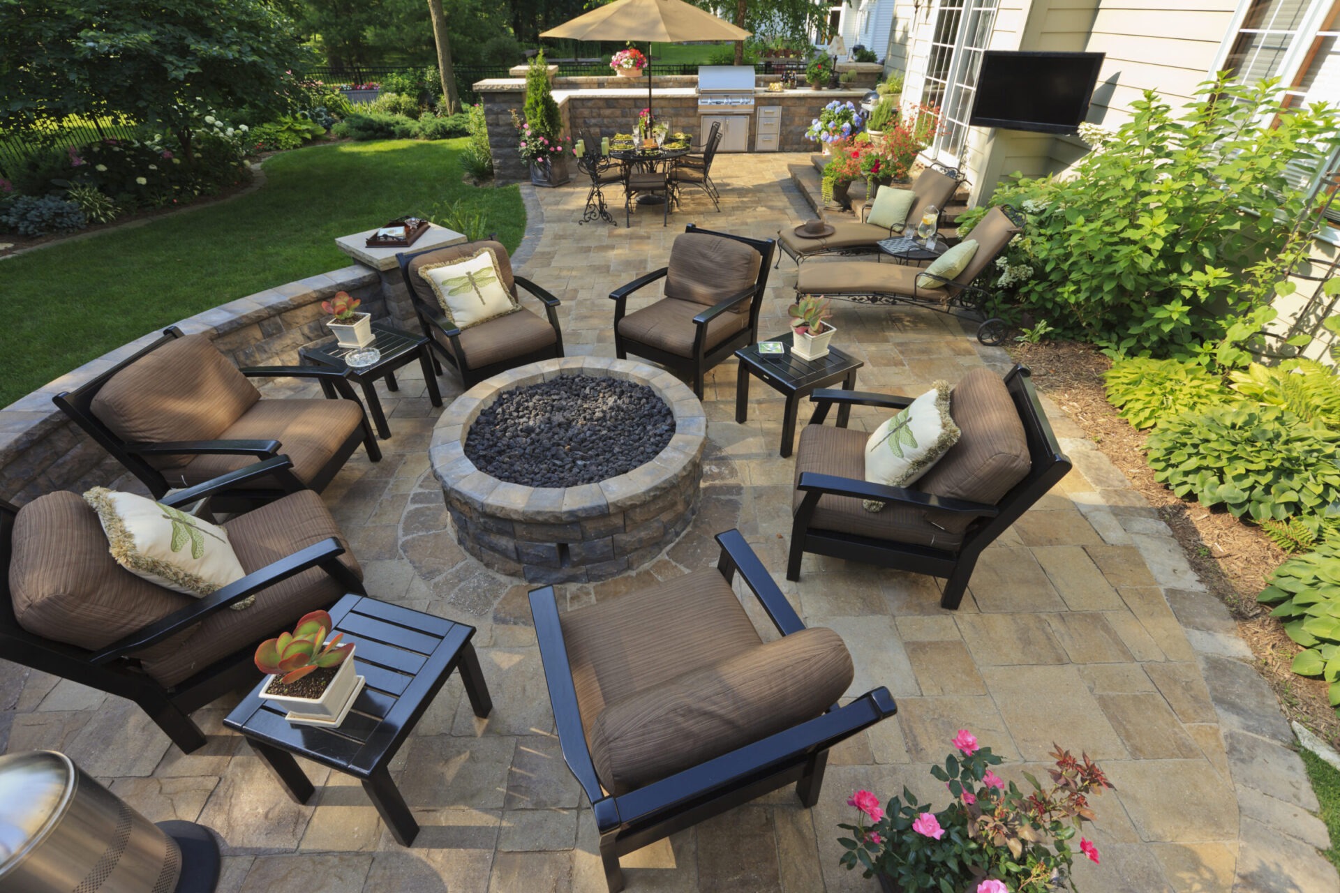 Cozy outdoor patio with stone fire pit, cushioned chairs, surrounding greenery, and dining area under an umbrella, perfect for relaxation and entertaining.