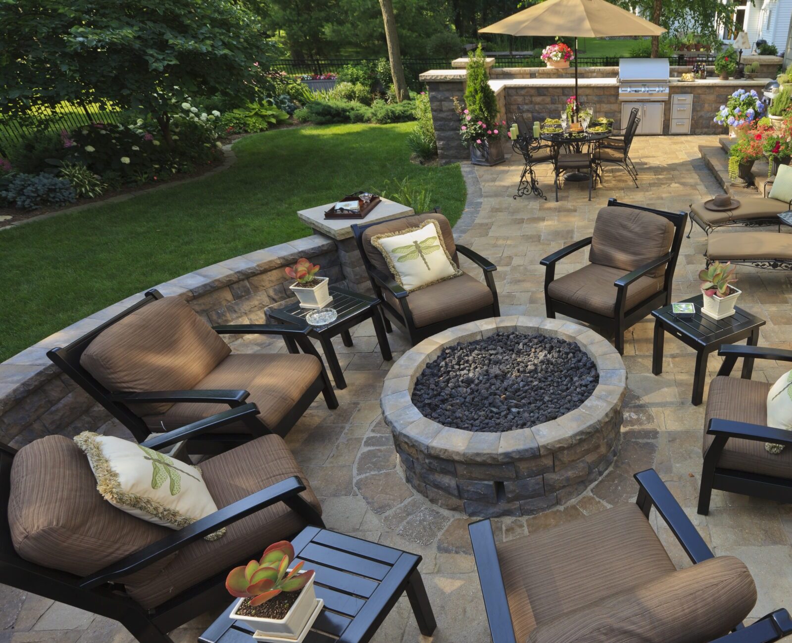 Cozy outdoor patio with stone fire pit, cushioned chairs, surrounding greenery, and dining area under an umbrella, perfect for relaxation and entertaining.