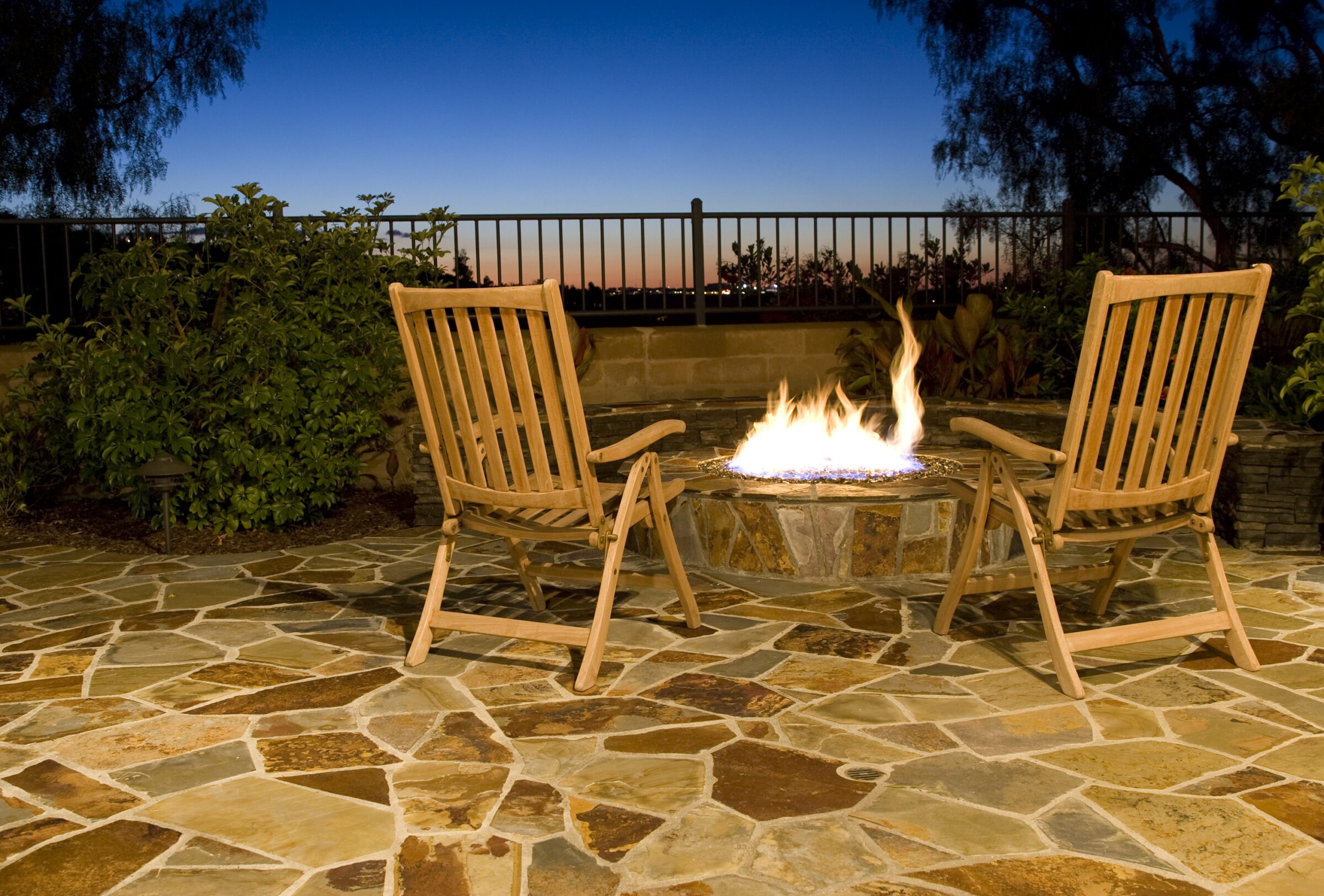 Two wooden chairs face a lit fire pit on a stone patio with a sunset sky and garden backdrop.