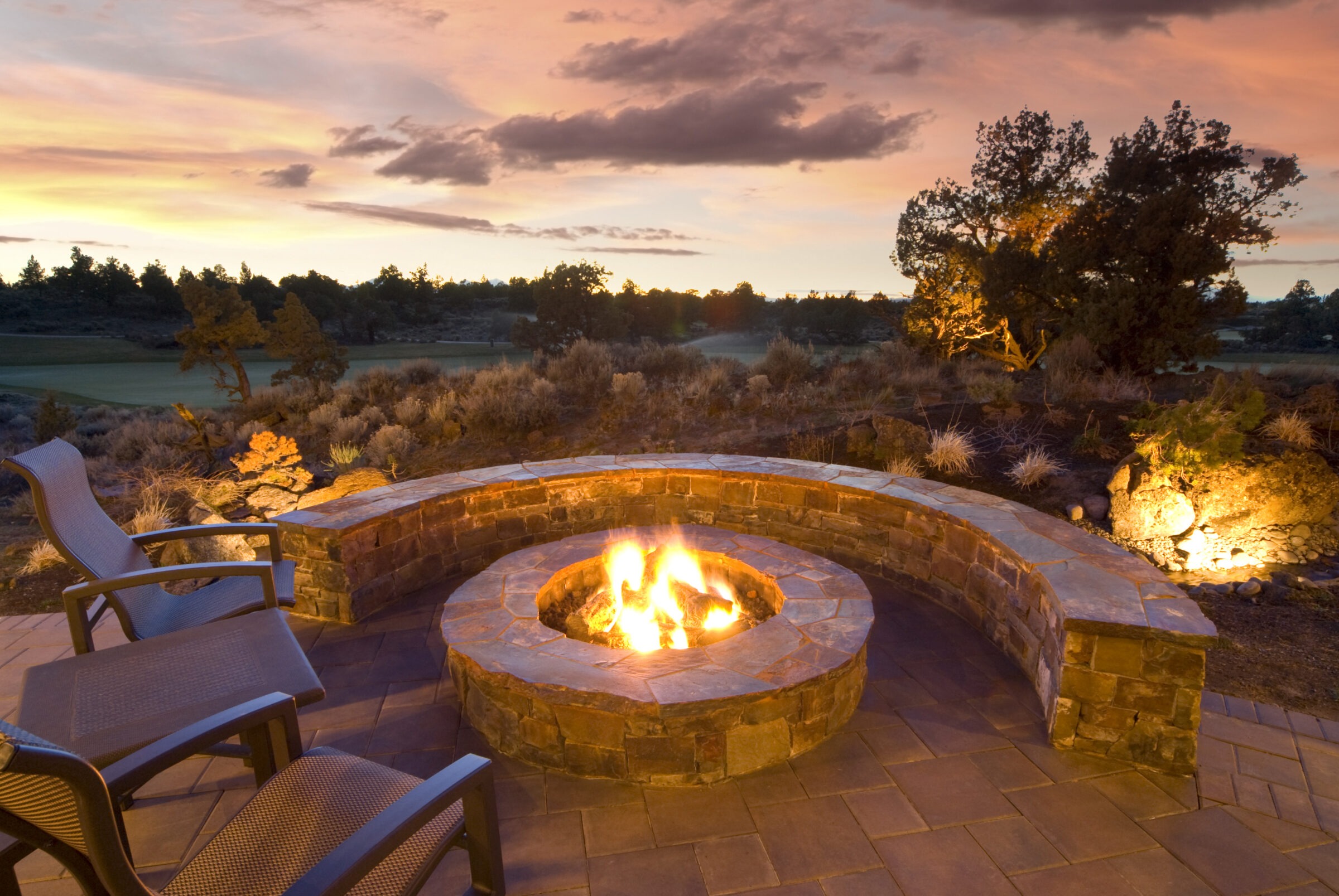 A stone fire pit surrounded by chairs and a curved bench at sunset; tranquil landscape with trees in the background.
