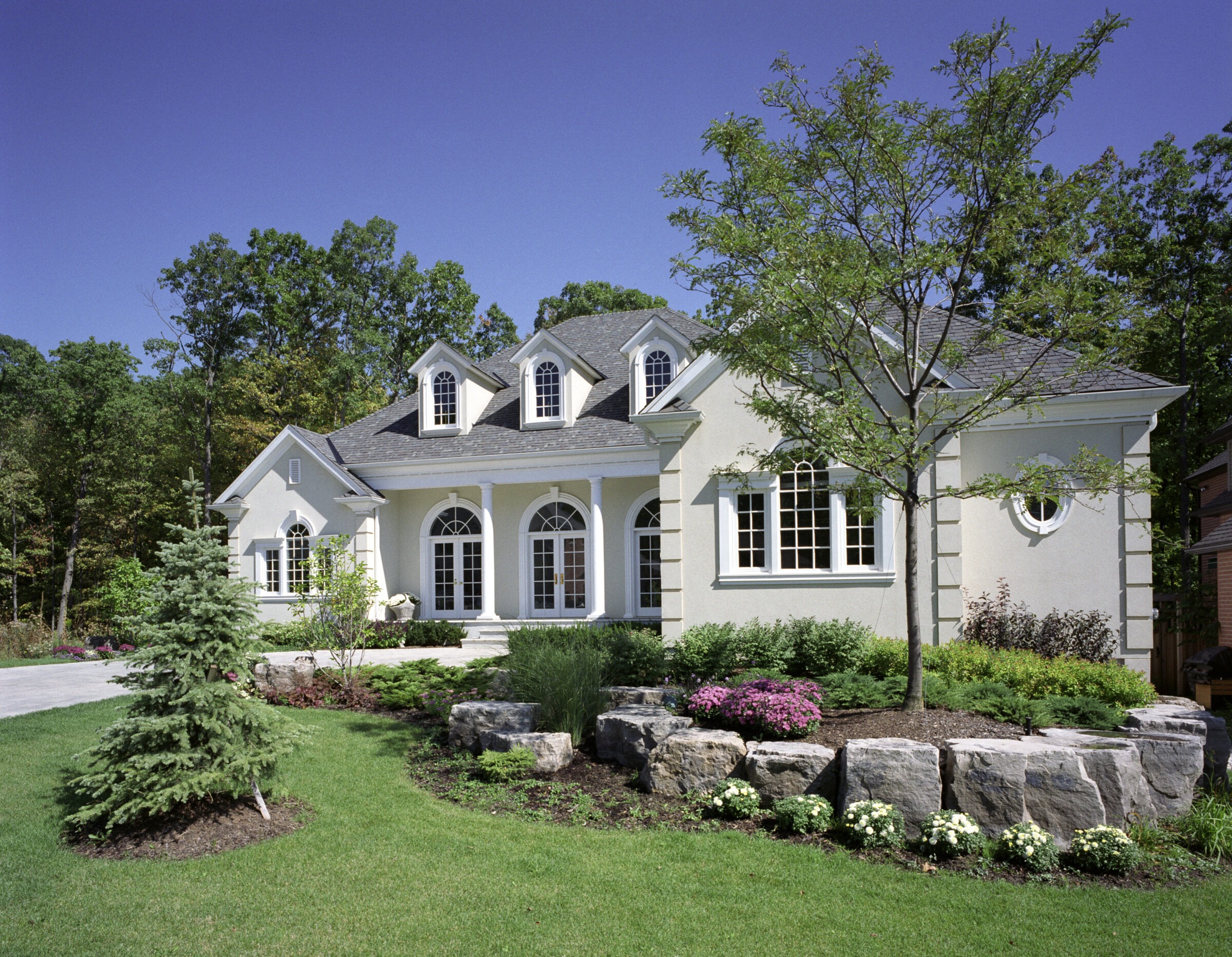 Elegant house with arched windows, surrounded by lush greenery and flowers. A tree and rock garden enhance the landscaped front yard.