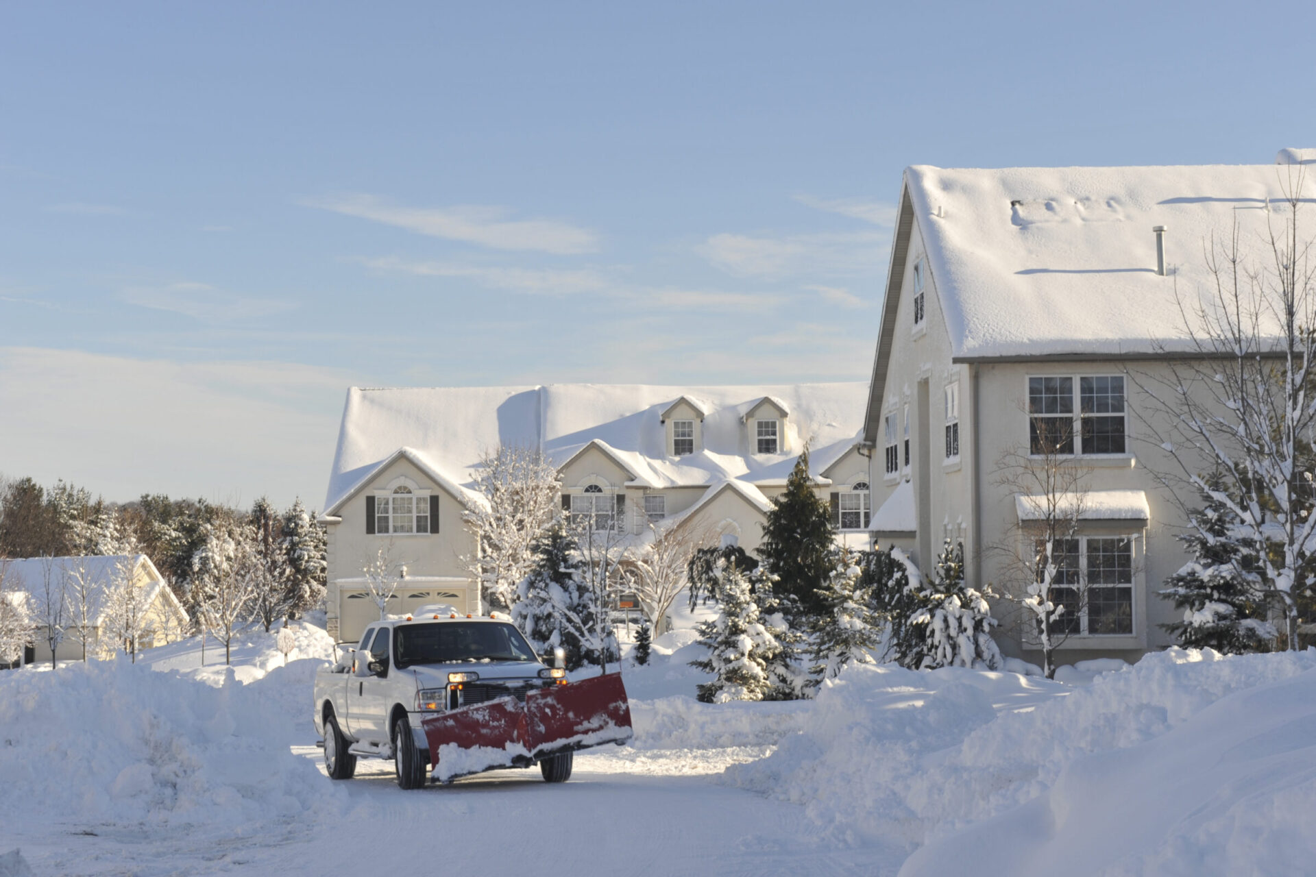 A snow-covered suburban neighborhood with a truck plowing snow on a driveway, surrounded by trees and houses under clear blue sky.