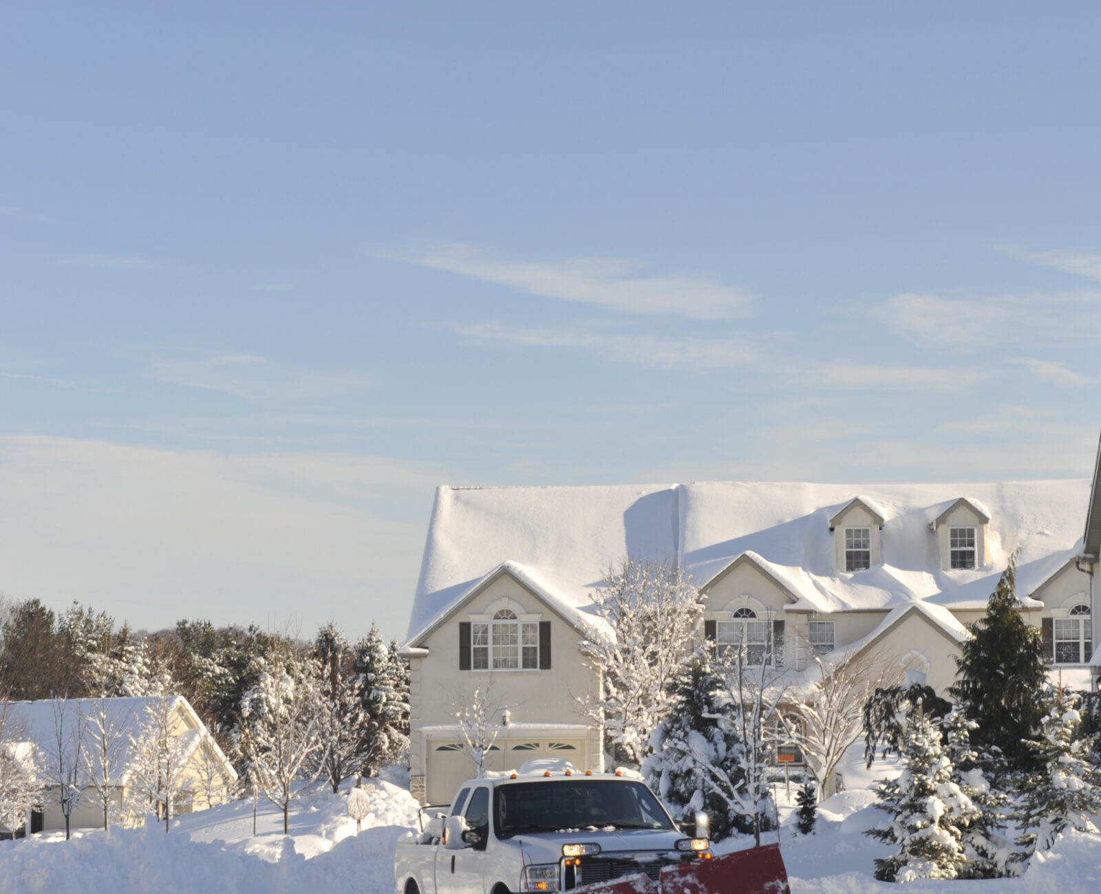 A snow-covered suburban neighborhood with a truck plowing snow on a driveway, surrounded by trees and houses under clear blue sky.
