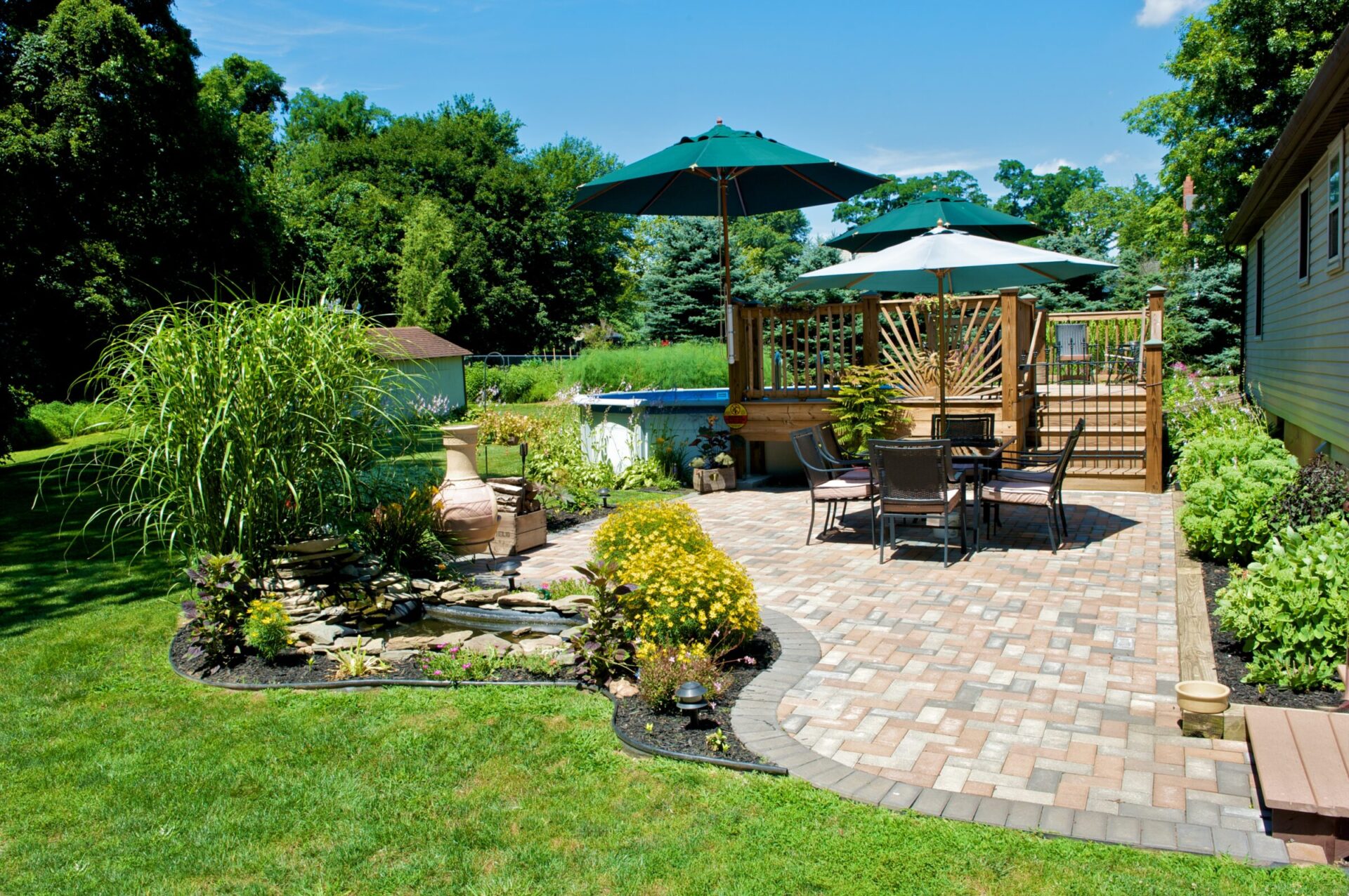 A sunny backyard features a patio with seating, umbrellas, lush greenery, and a small garden with a pond, amidst vibrant landscaping.
