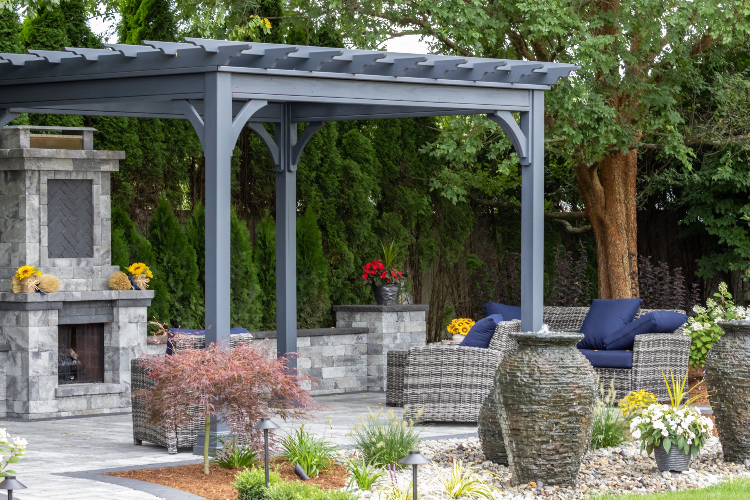 A beautifully landscaped patio features a pergola, stone fireplace, wicker furniture with blue cushions, vibrant plants, and a serene outdoor atmosphere.