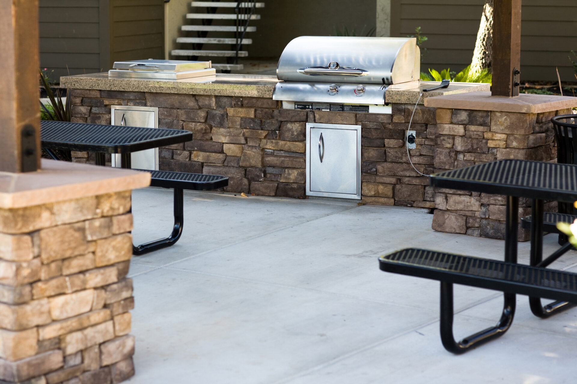 Outdoor grilling area with stone countertops and built-in grill, surrounded by black metal picnic tables on concrete patio, under a shaded structure.