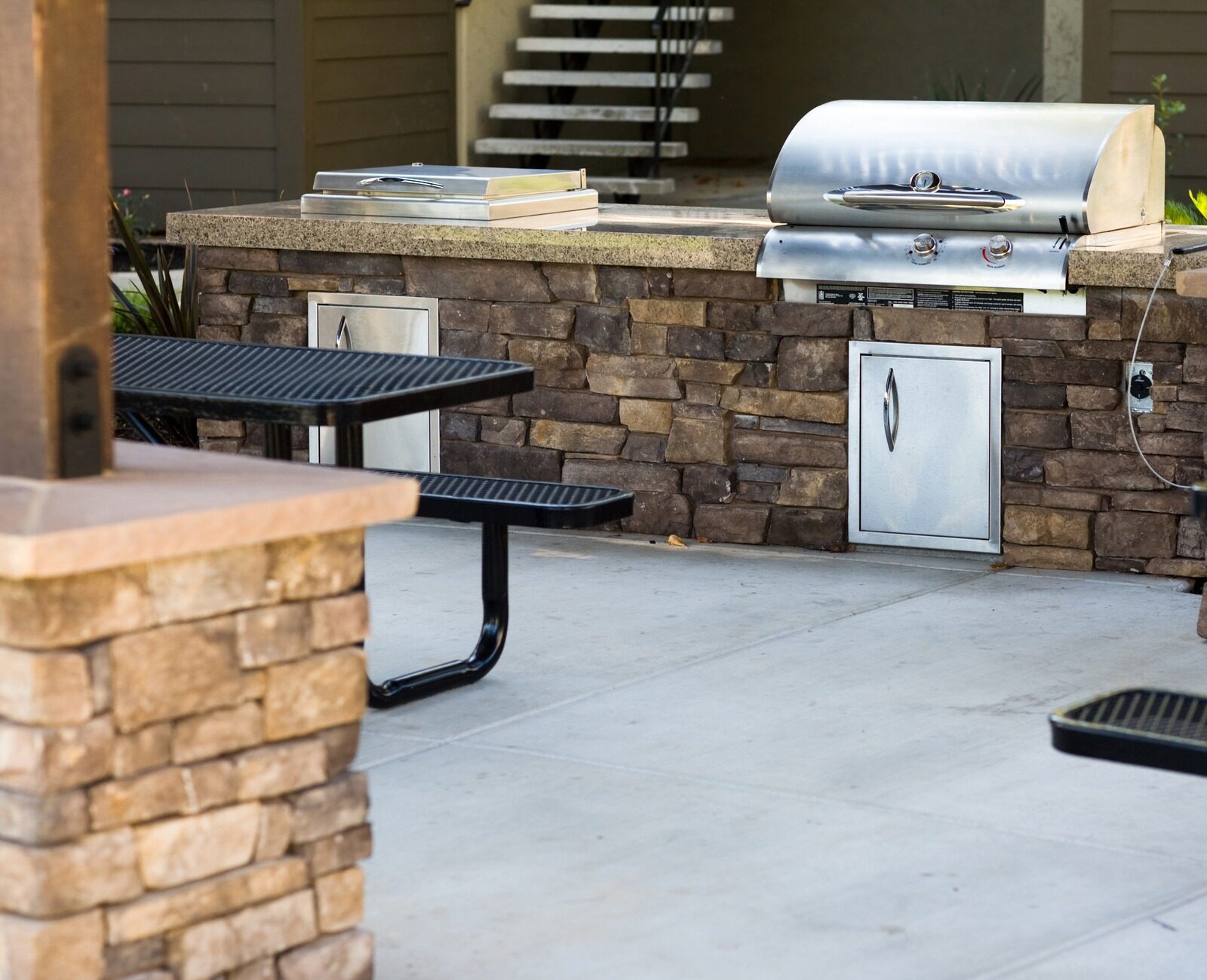 Outdoor grilling area with stone countertops and built-in grill, surrounded by black metal picnic tables on concrete patio, under a shaded structure.