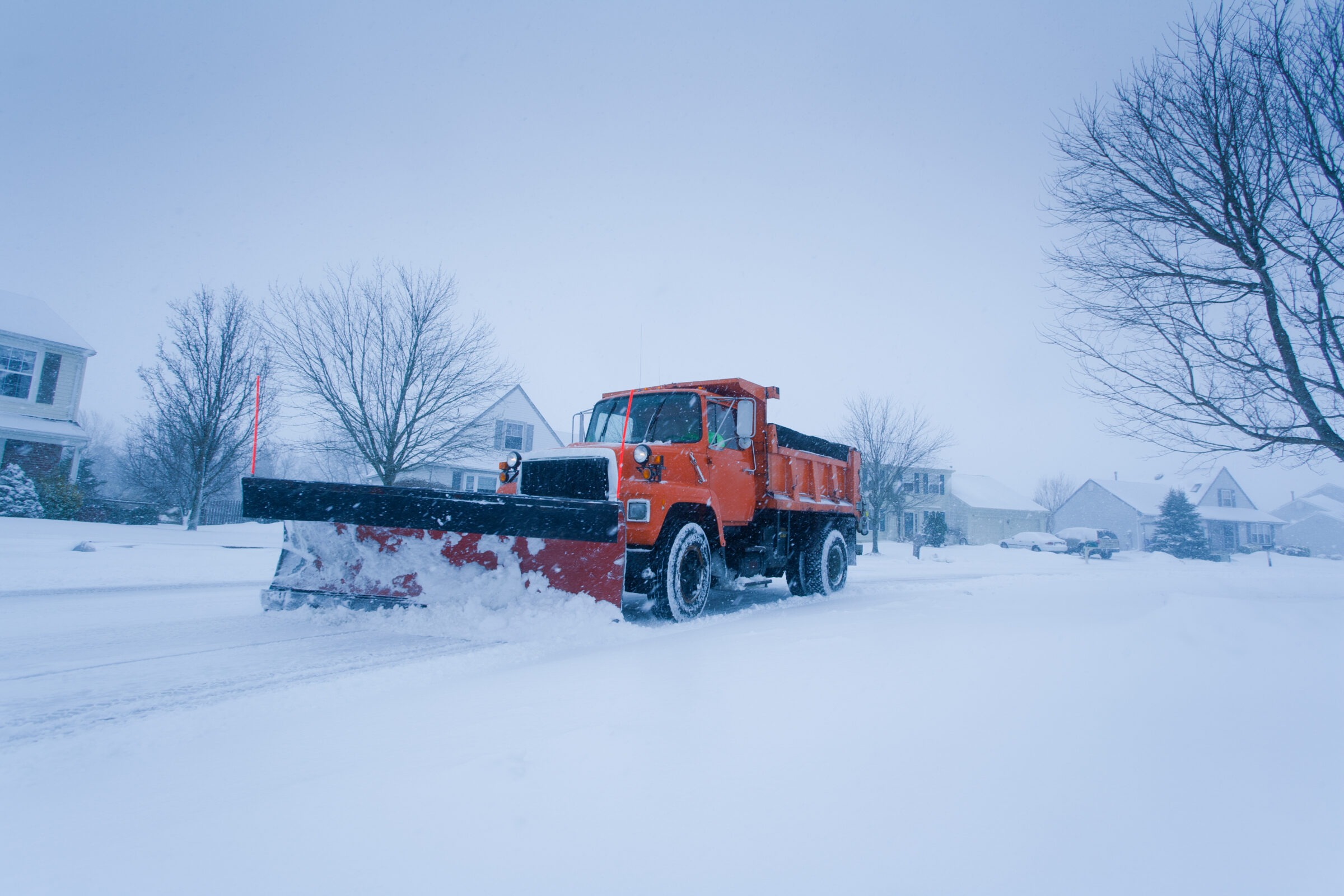 An orange snowplow truck clears a snowy suburban street lined with houses and bare trees, creating a path through the winter landscape.