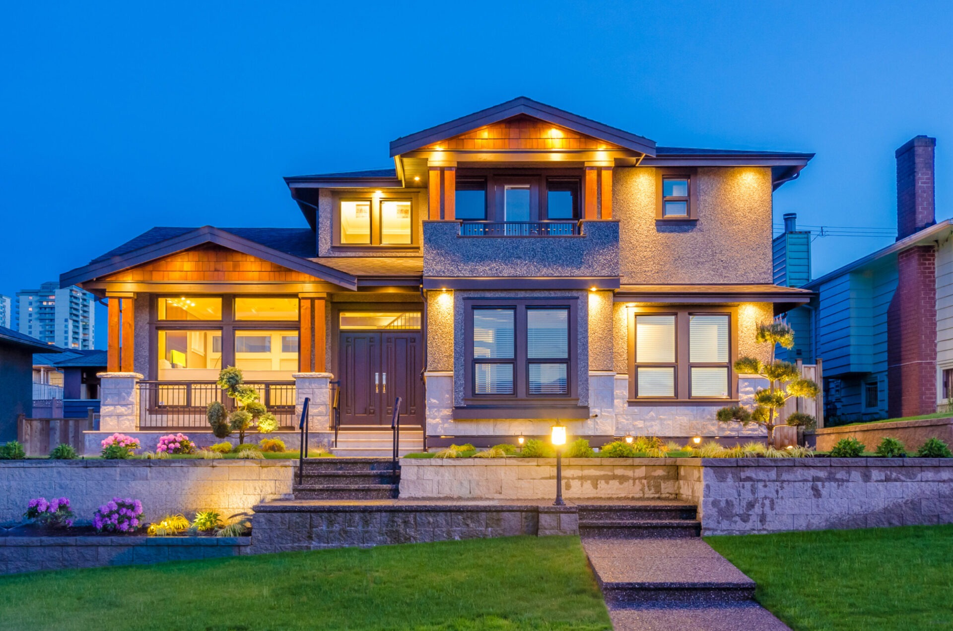A modern two-story house with illuminated exterior and manicured garden. Urban buildings in the background under a twilight sky.