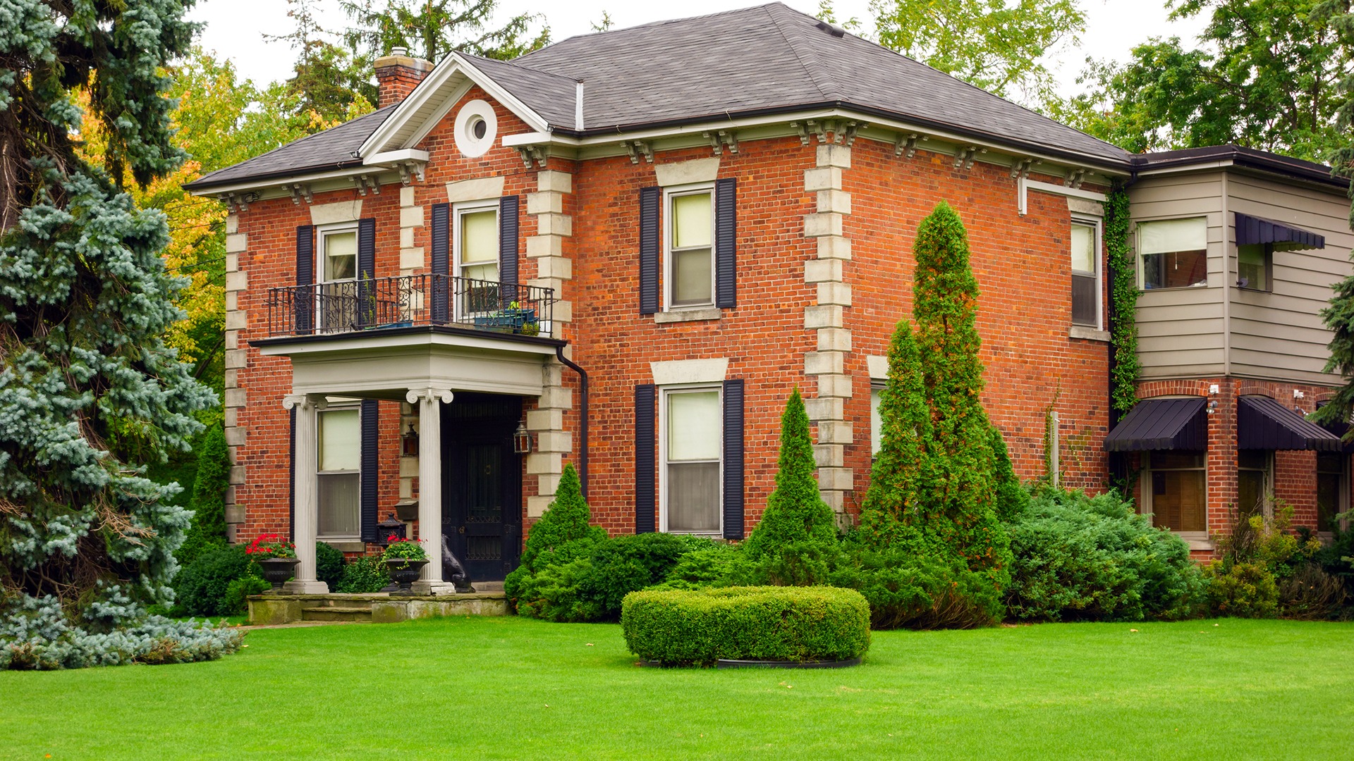 A stately red brick house with a well-maintained garden, tall trees, and a small balcony, showcasing classic architectural details.