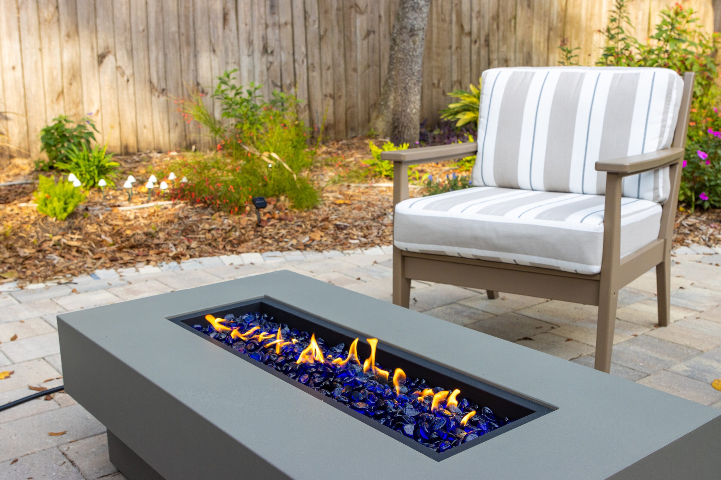 Patio scene with striped cushioned chair beside a modern fire pit, featuring blue glass pebbles and orange flames, surrounded by greenery and fence.